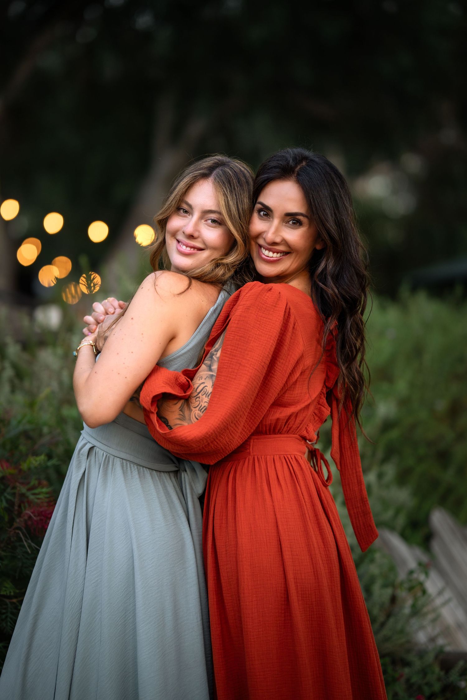Two smiling women in elegant dresses embrace outdoors with warm bokeh lights in the background.