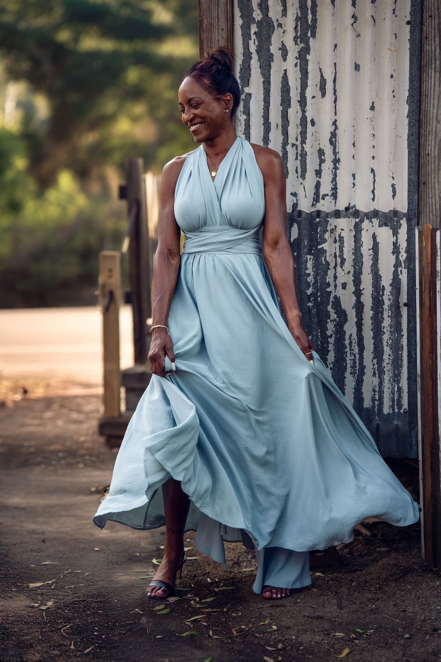 Woman in flowing light blue halter gown poses gracefully outdoors near rustic wall, smiling confidently.