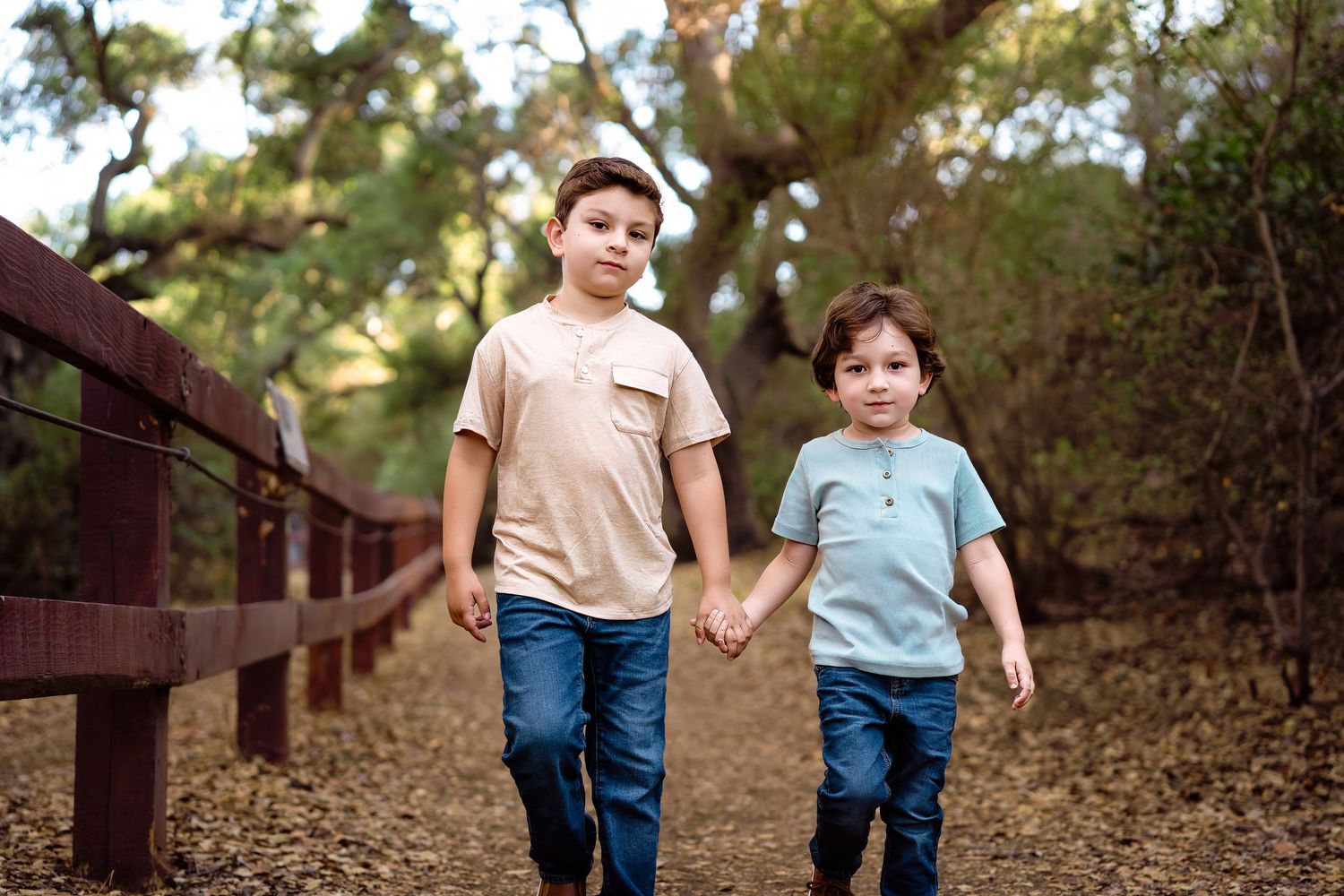 Two young children walk hand in hand along a wooden path through a wooded area in autumn.