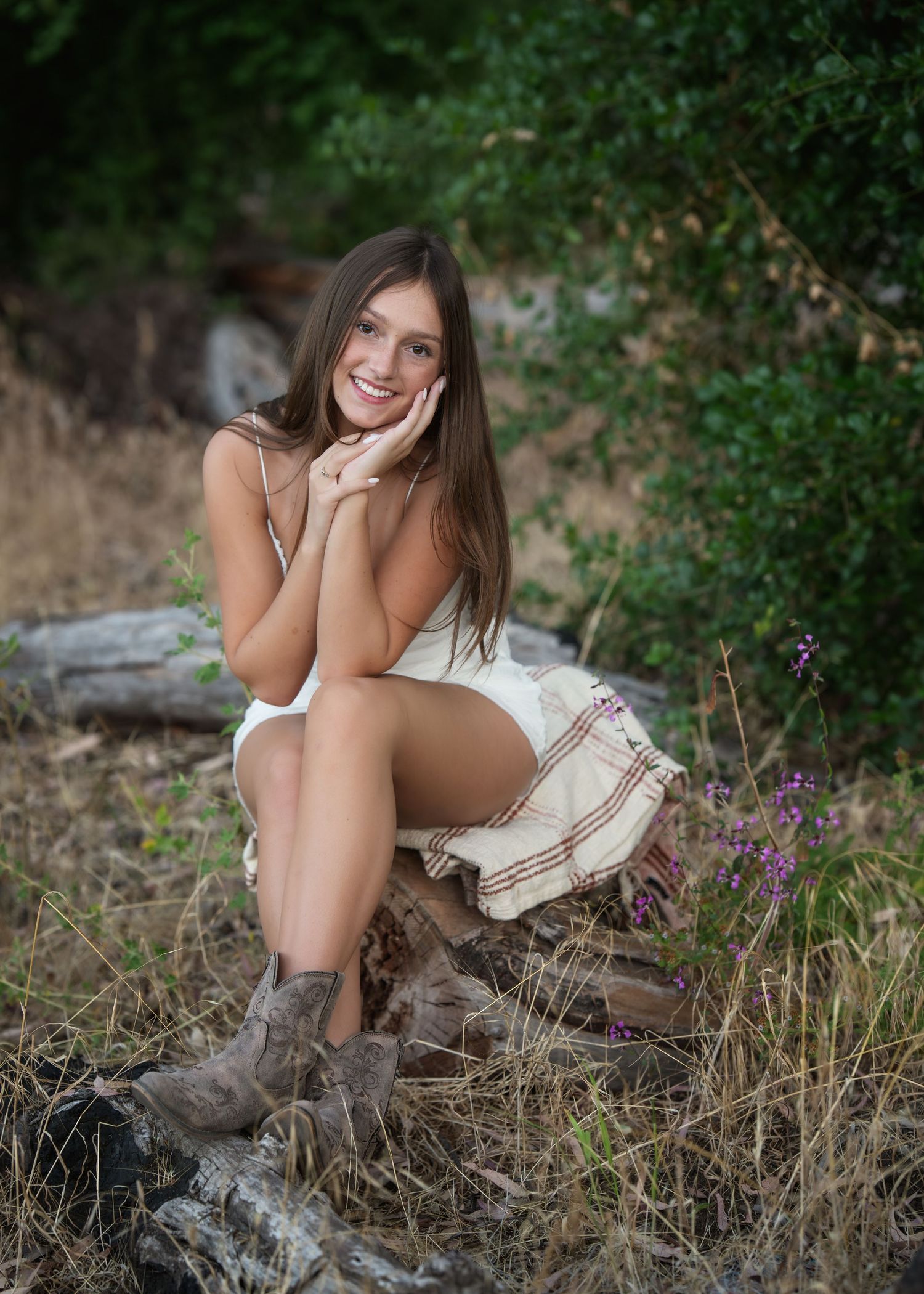 Young woman in white dress and brown boots sits smiling on a log in a natural outdoor setting.