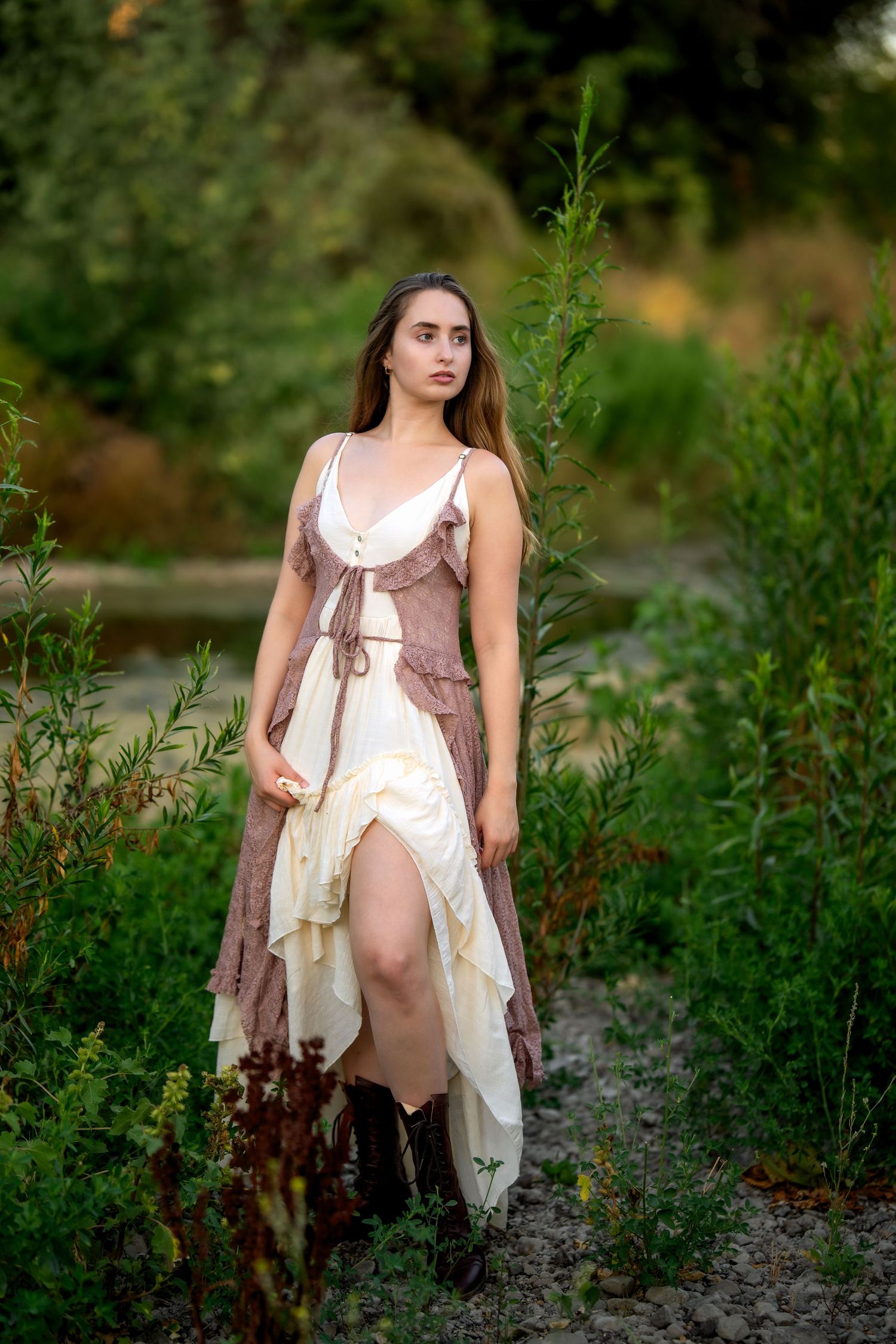 Young woman in bohemian layered dress and cowboy boots standing among lush green plants outdoors.