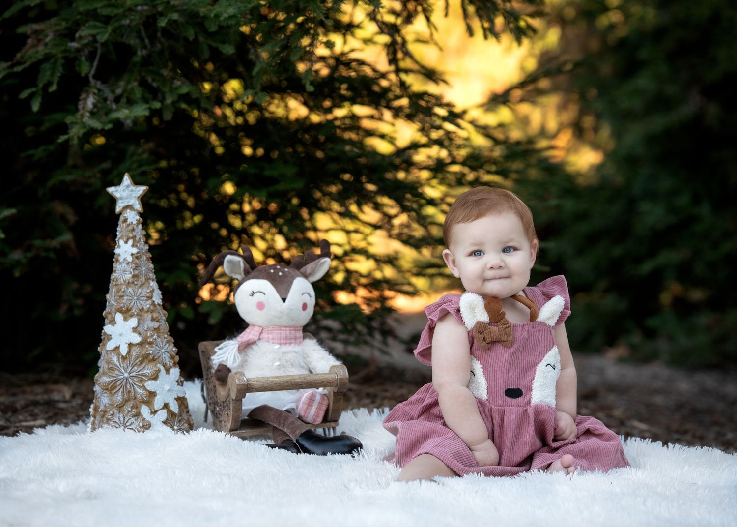 A winter scene with a small child in pink sitting in snow beside a decorative Christmas tree and plush bunny at sunset.