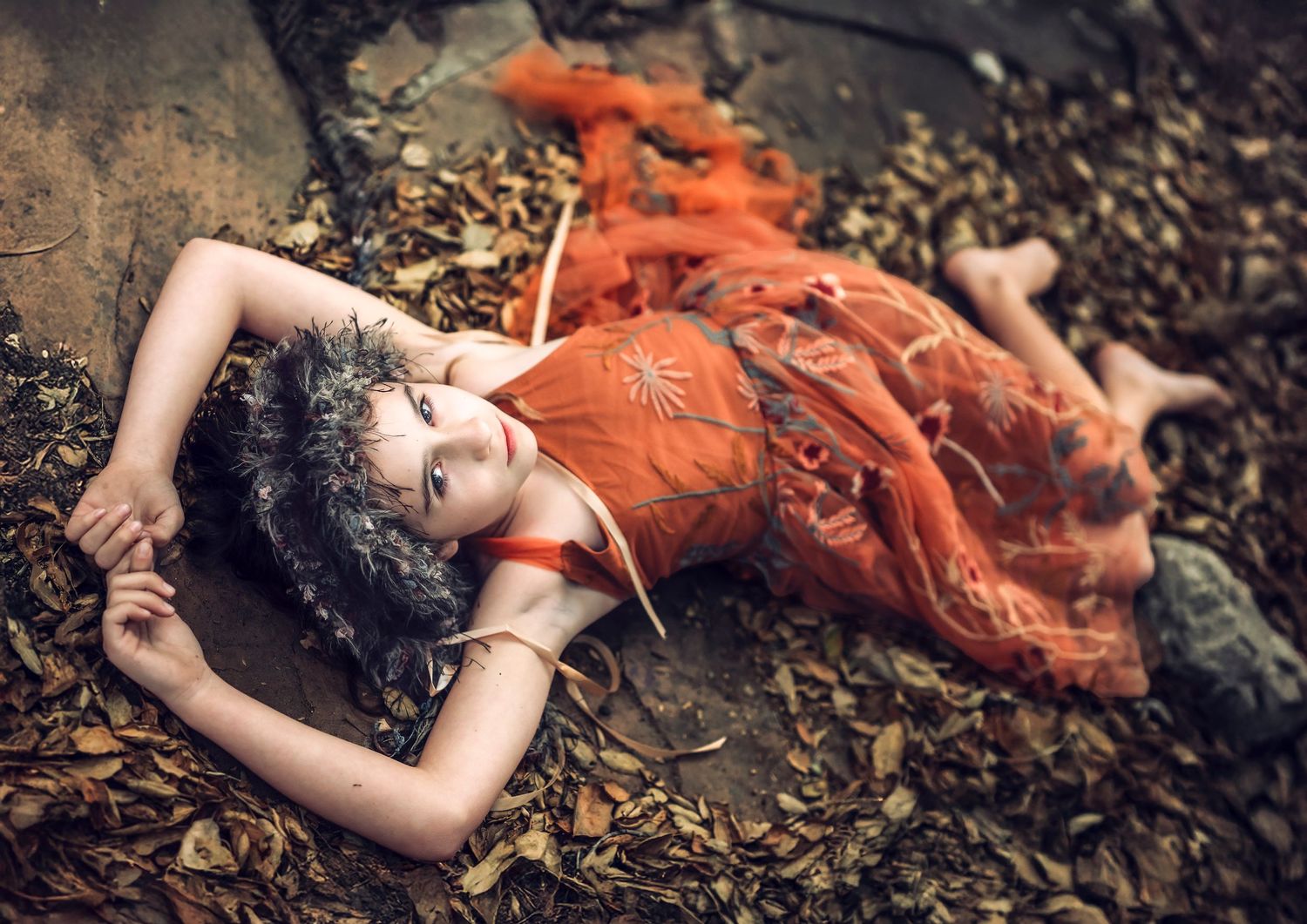 Woman in orange dress lying artistically on forest floor with scattered leaves.