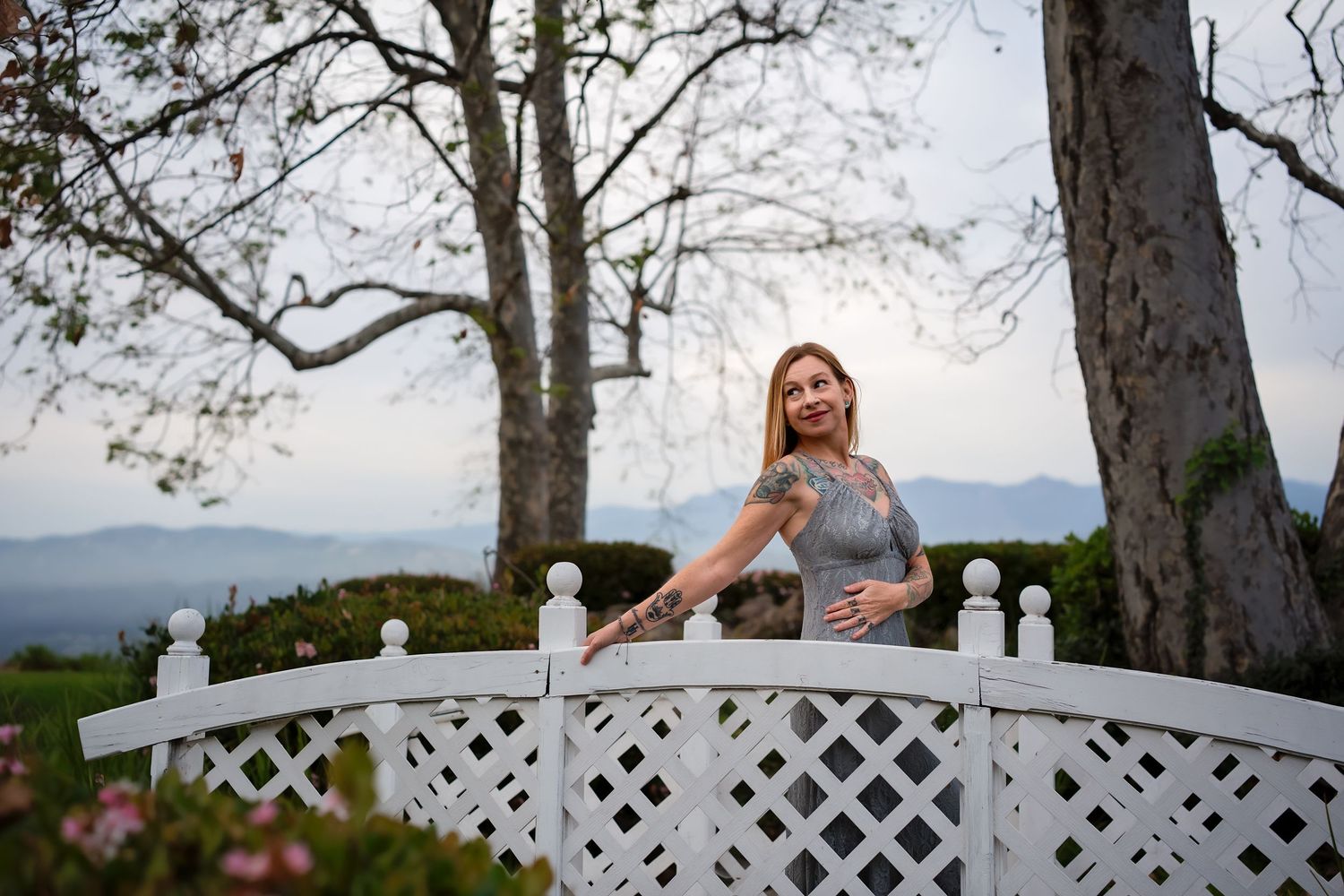 Woman in gray dress leans on white lattice bridge surrounded by trees and mountain views.