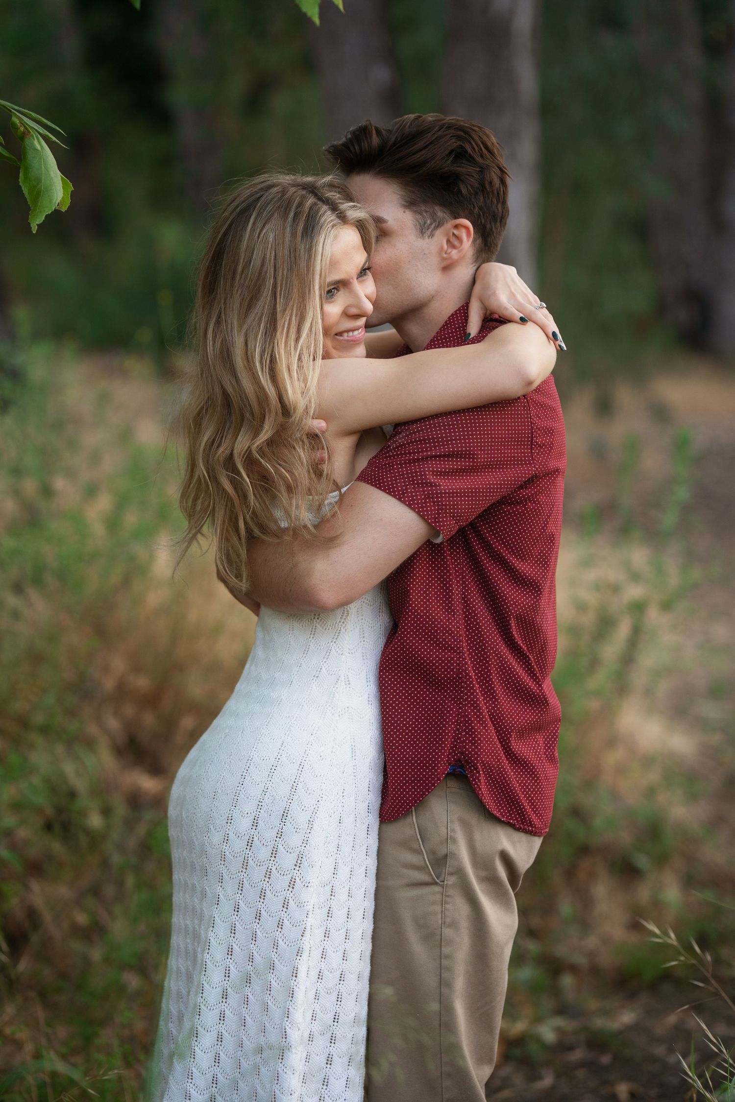 A couple embraces in a forest, woman in white dress, man in red shirt, sharing a tender romantic moment outdoors.