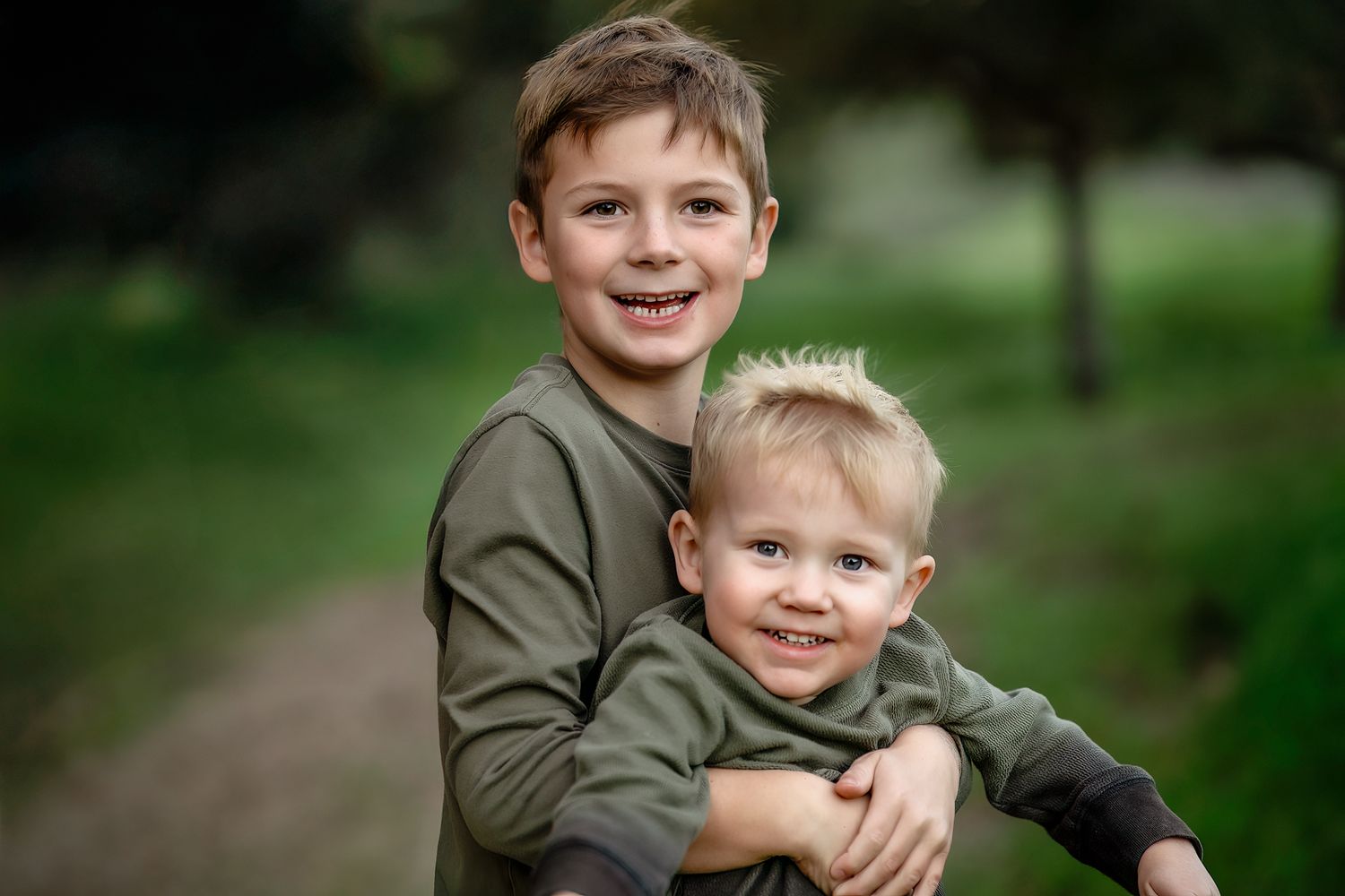 Two young boys in olive green shirts share a warm embrace while smiling at the camera outdoors.