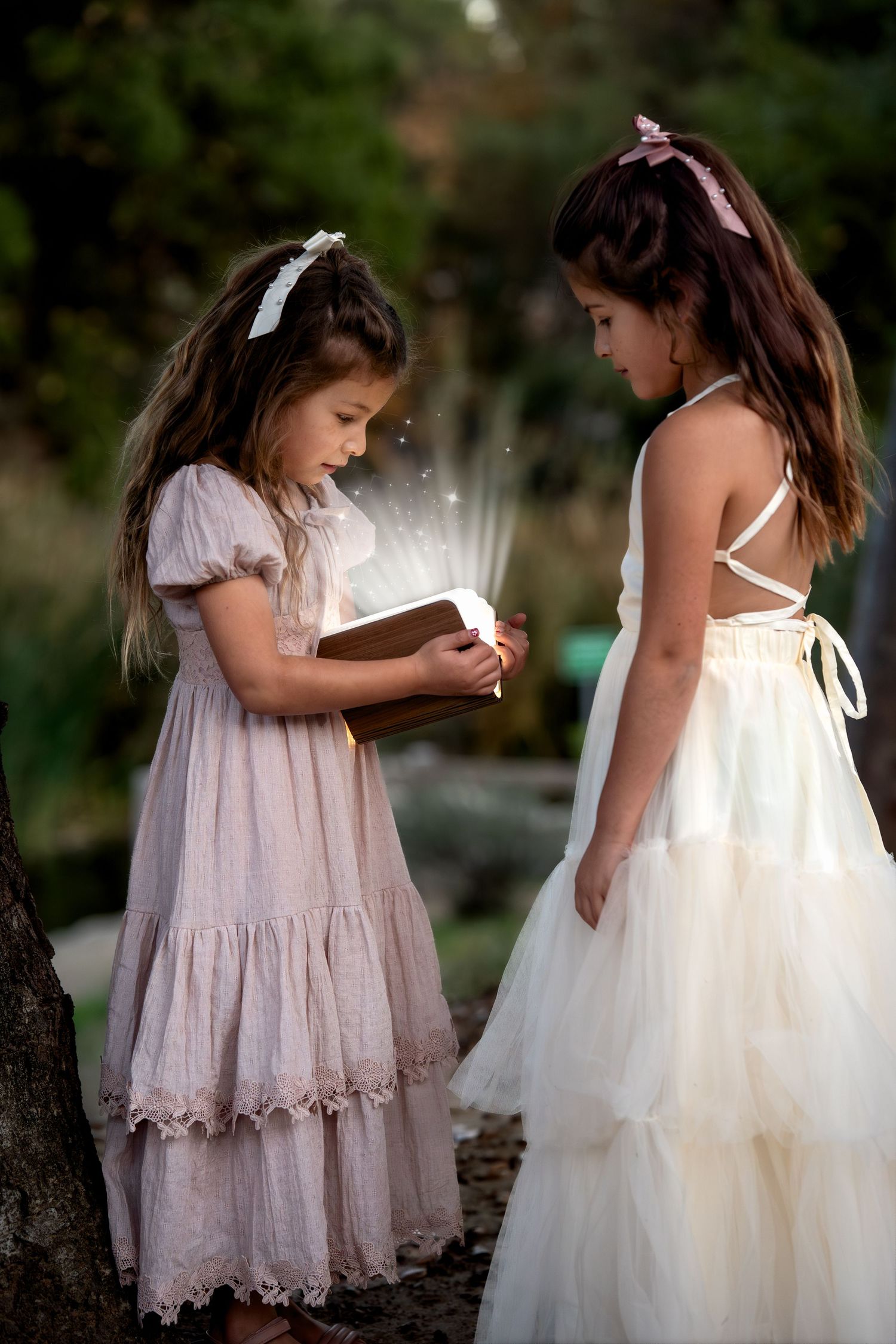 Two young girls in flowing dresses stand together outdoors, one reading from a small book in a dreamy natural setting.