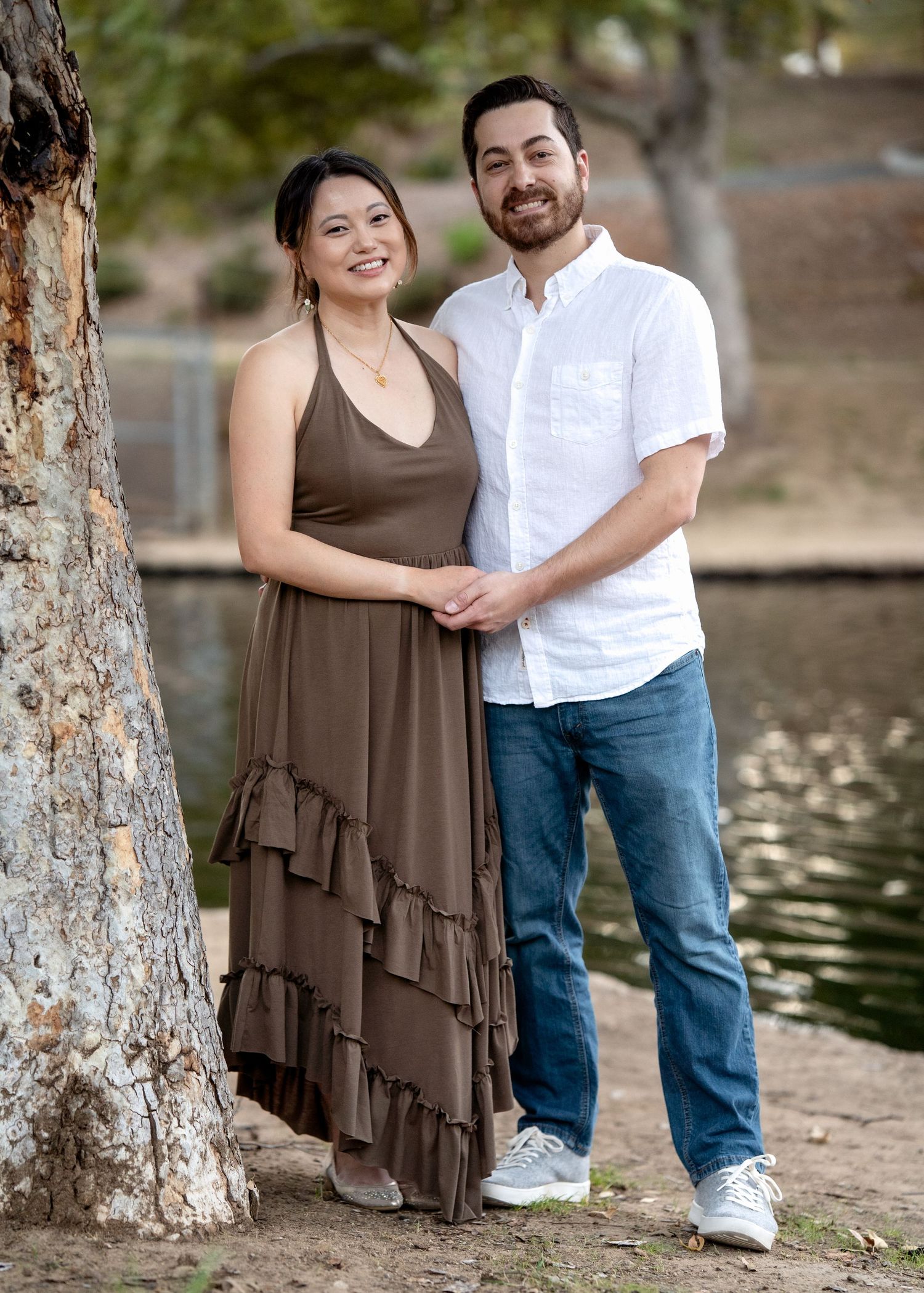 Couple posing casually by tree-lined pond in coordinated outfits.