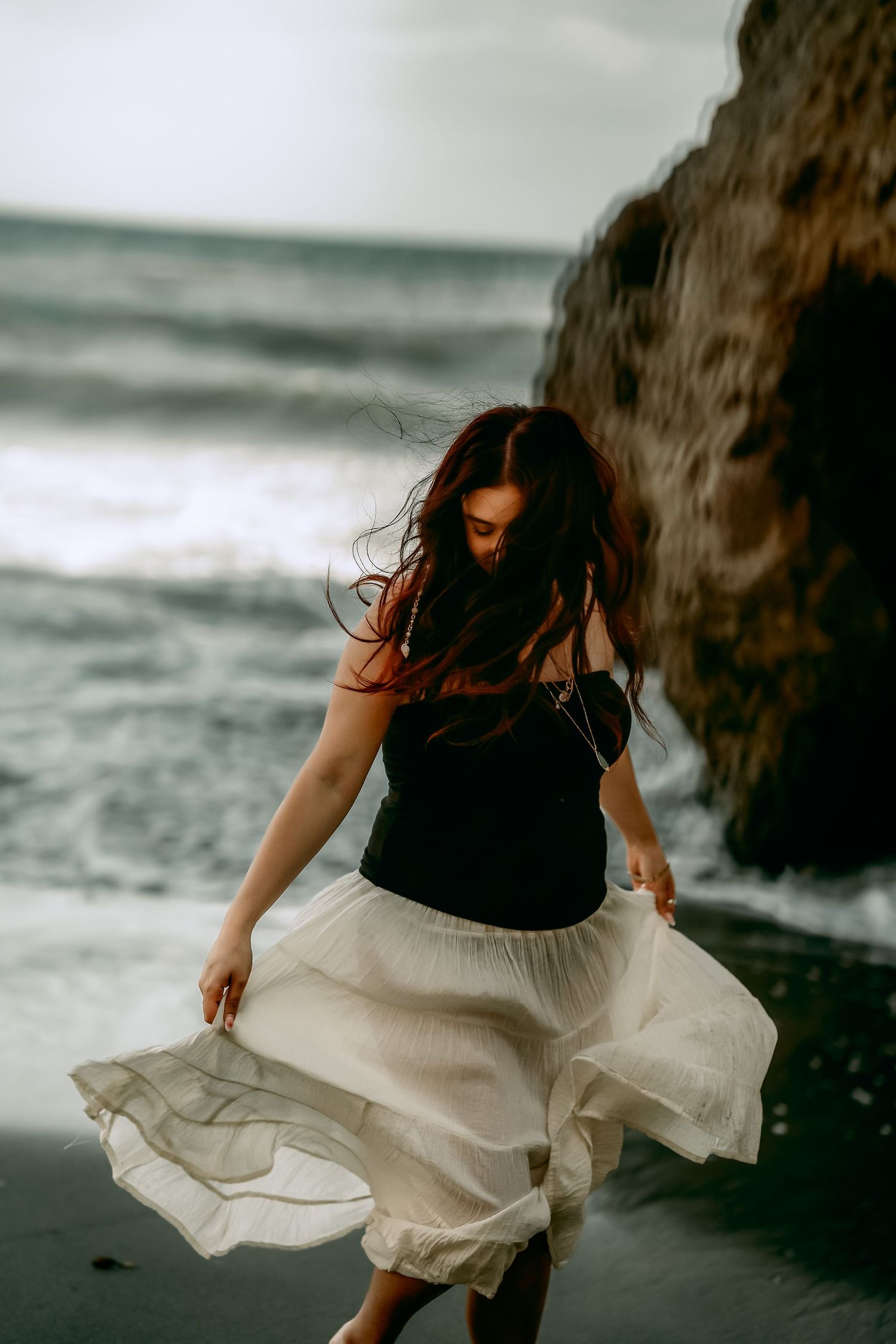 Figure in flowing white skirt dancing on moody beach with crashing waves.