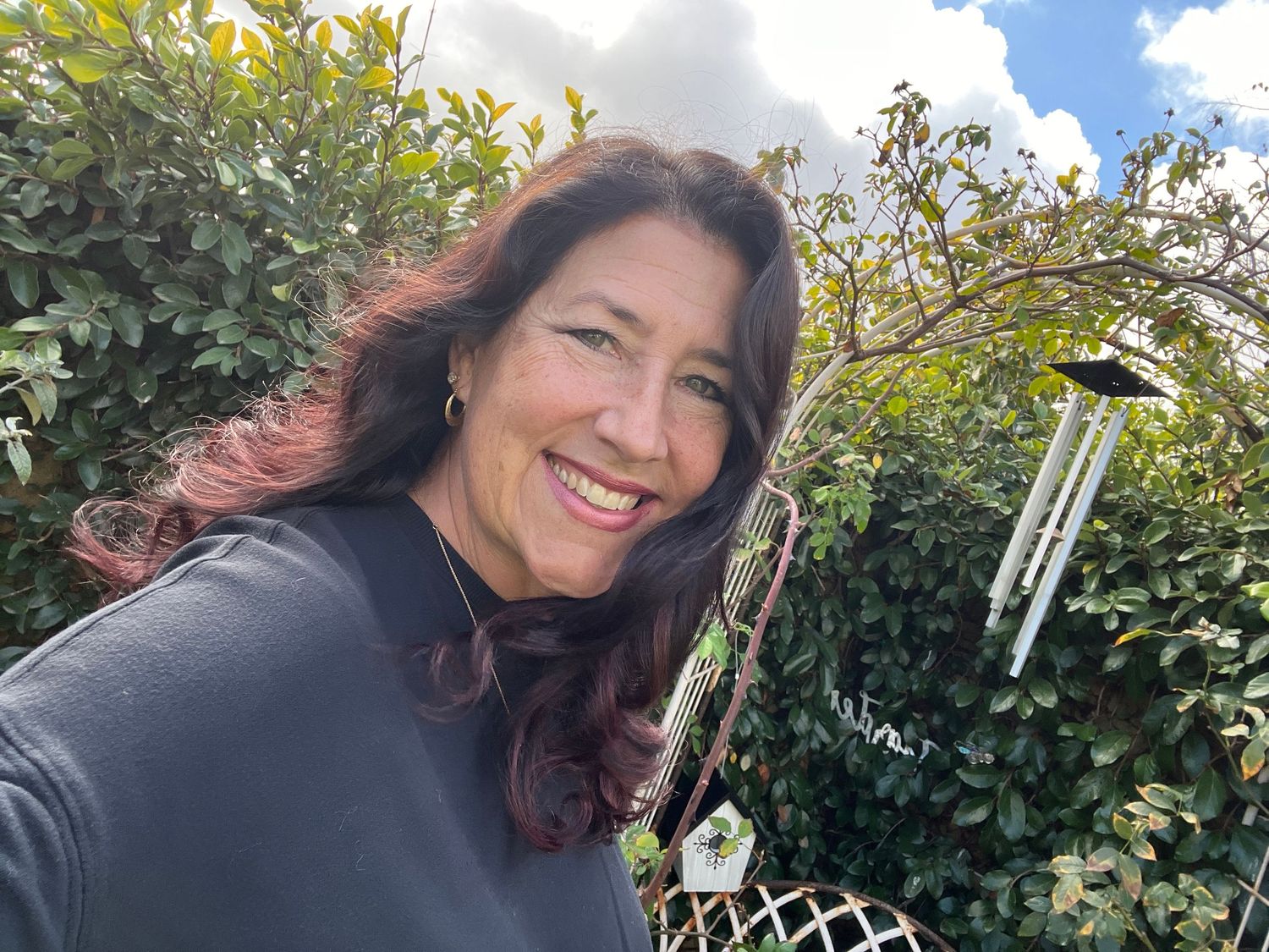 Selfie outdoors with wind chimes and greenery visible in background.