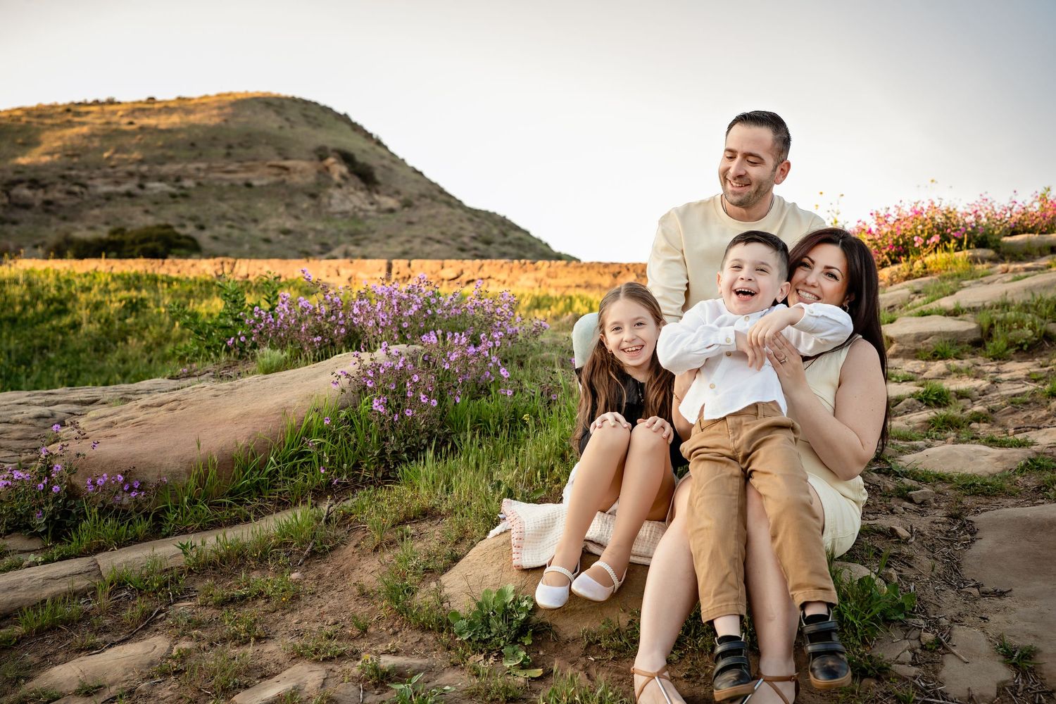 Couple sitting on rocky terrain with wildflowers during golden hour sunset in a mountain landscape.