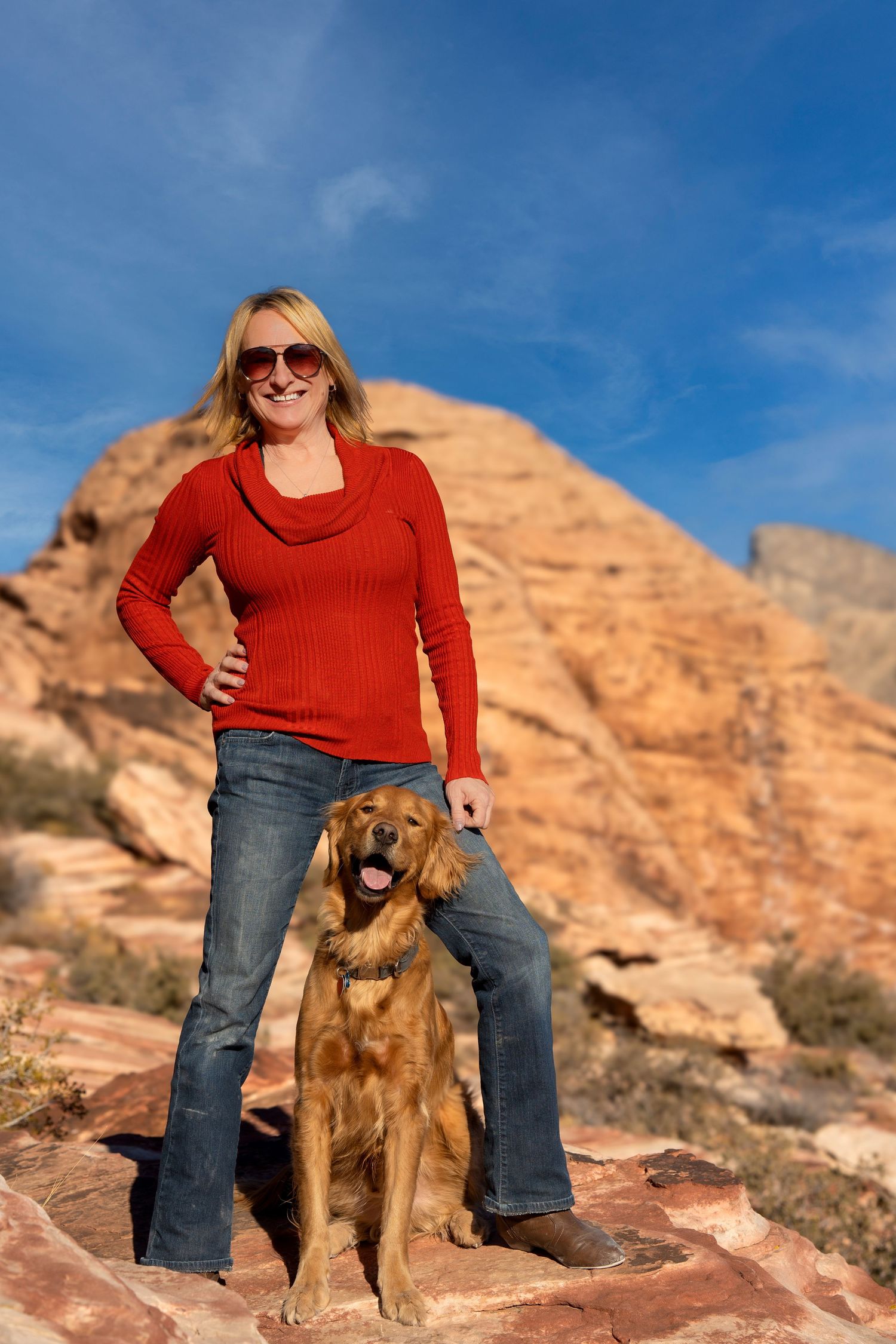 A blonde woman in a red sweater and jeans poses with her golden Irish Setter dog on red rocks with blue sky.