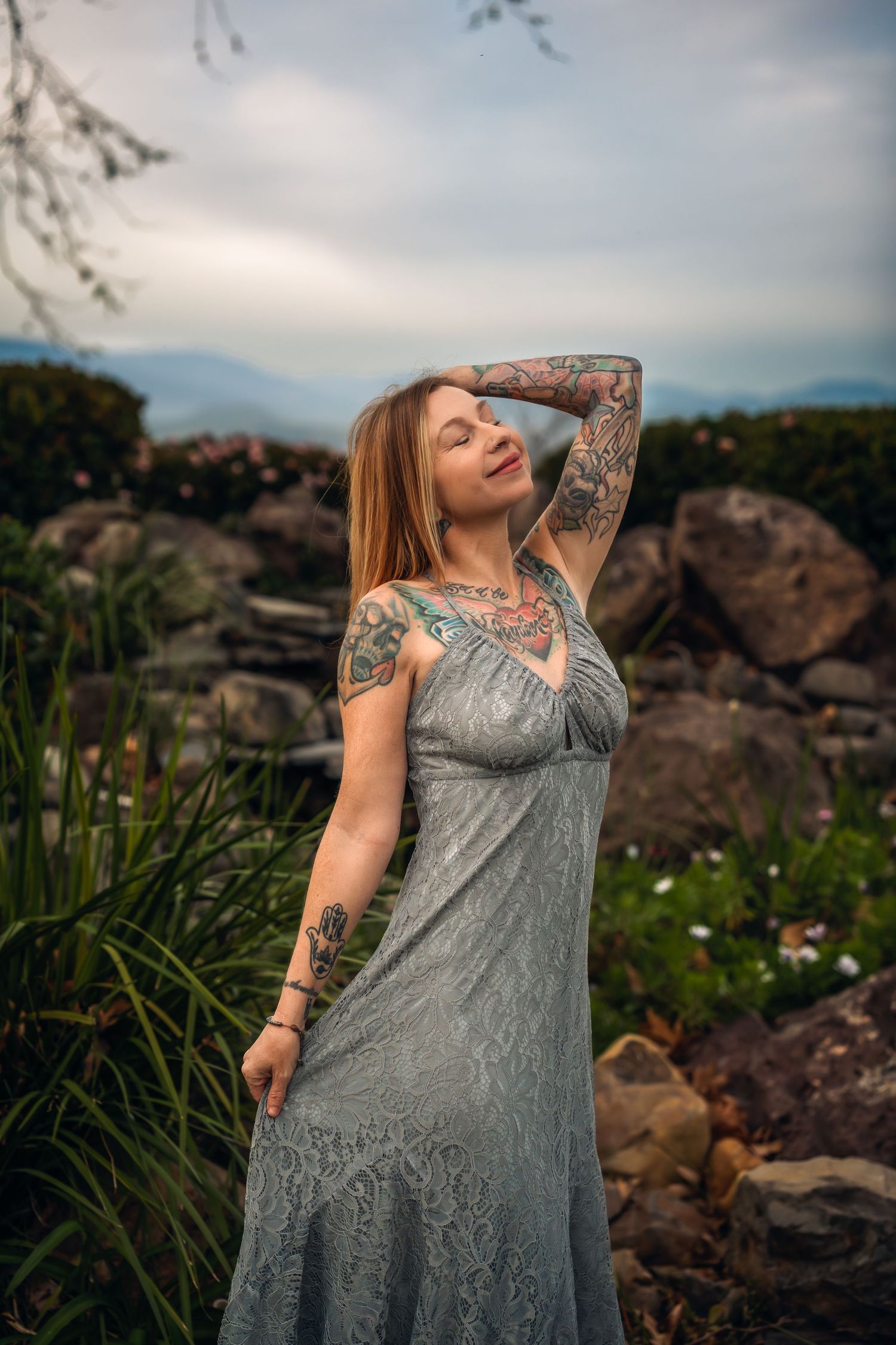 Tattooed woman in gray lace dress poses gracefully outdoors among rocks and greenery with mountains in background.
