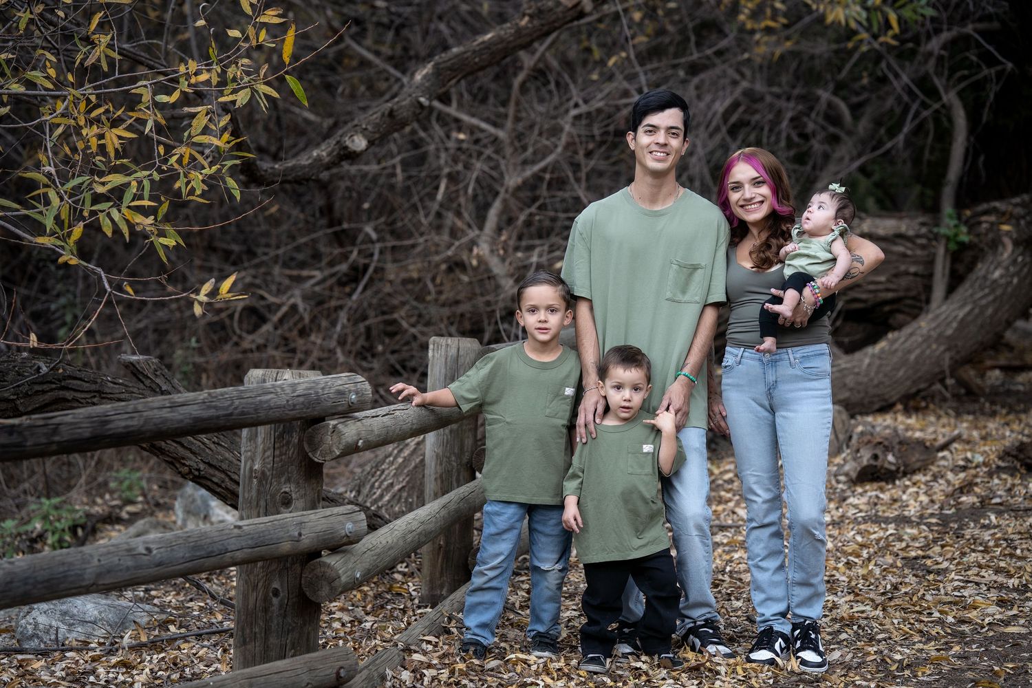A family in coordinating green shirts stands together by a rustic wooden fence in a wooded outdoor setting.