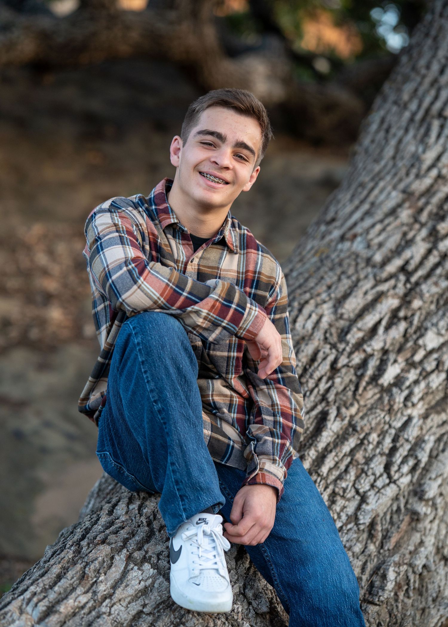 A person in a plaid flannel shirt and jeans sits casually on a tree trunk during an outdoor portrait session.