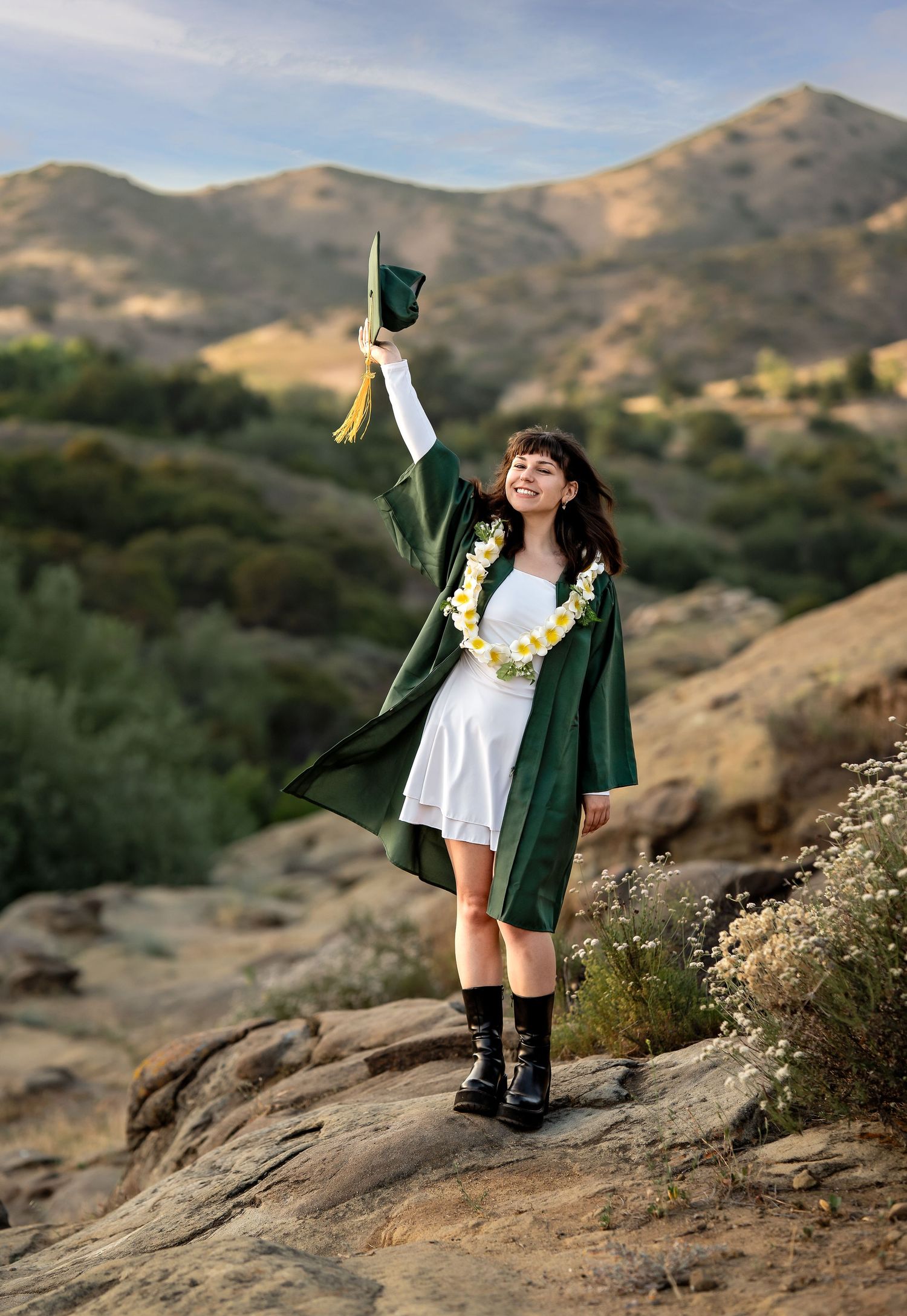 Graduate in dark green cap and gown celebrates on rocky mountain terrain.