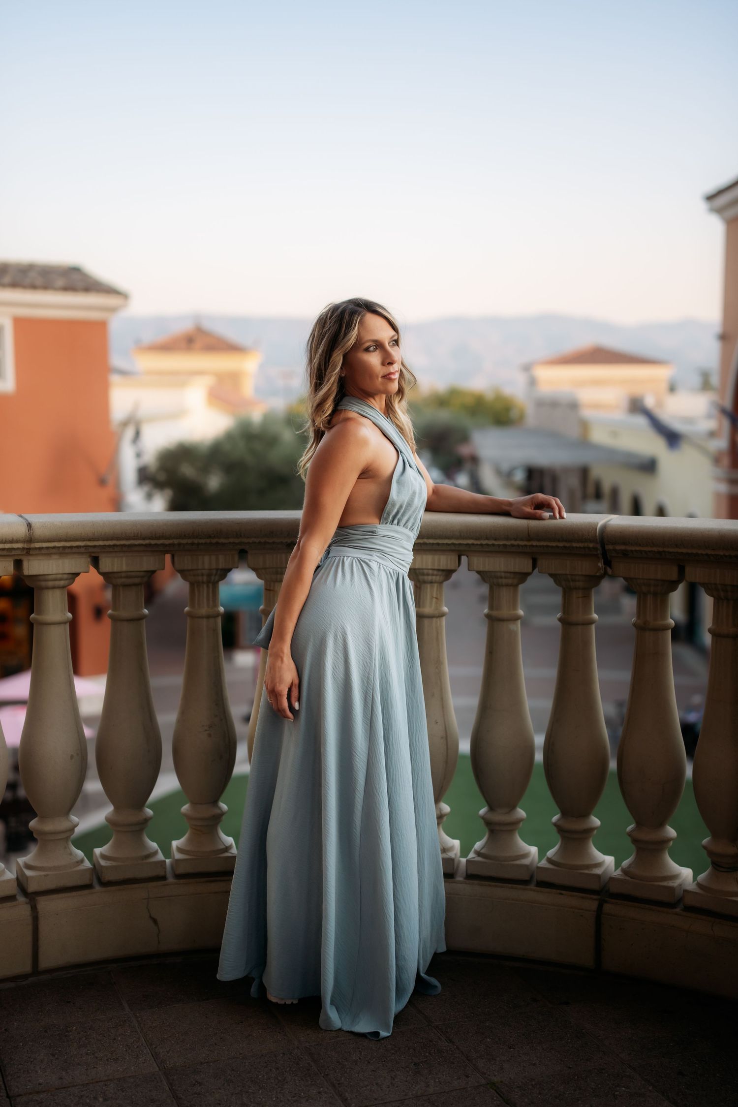 Woman in elegant light blue maxi dress posing on a balcony with balusters and scenic town views.