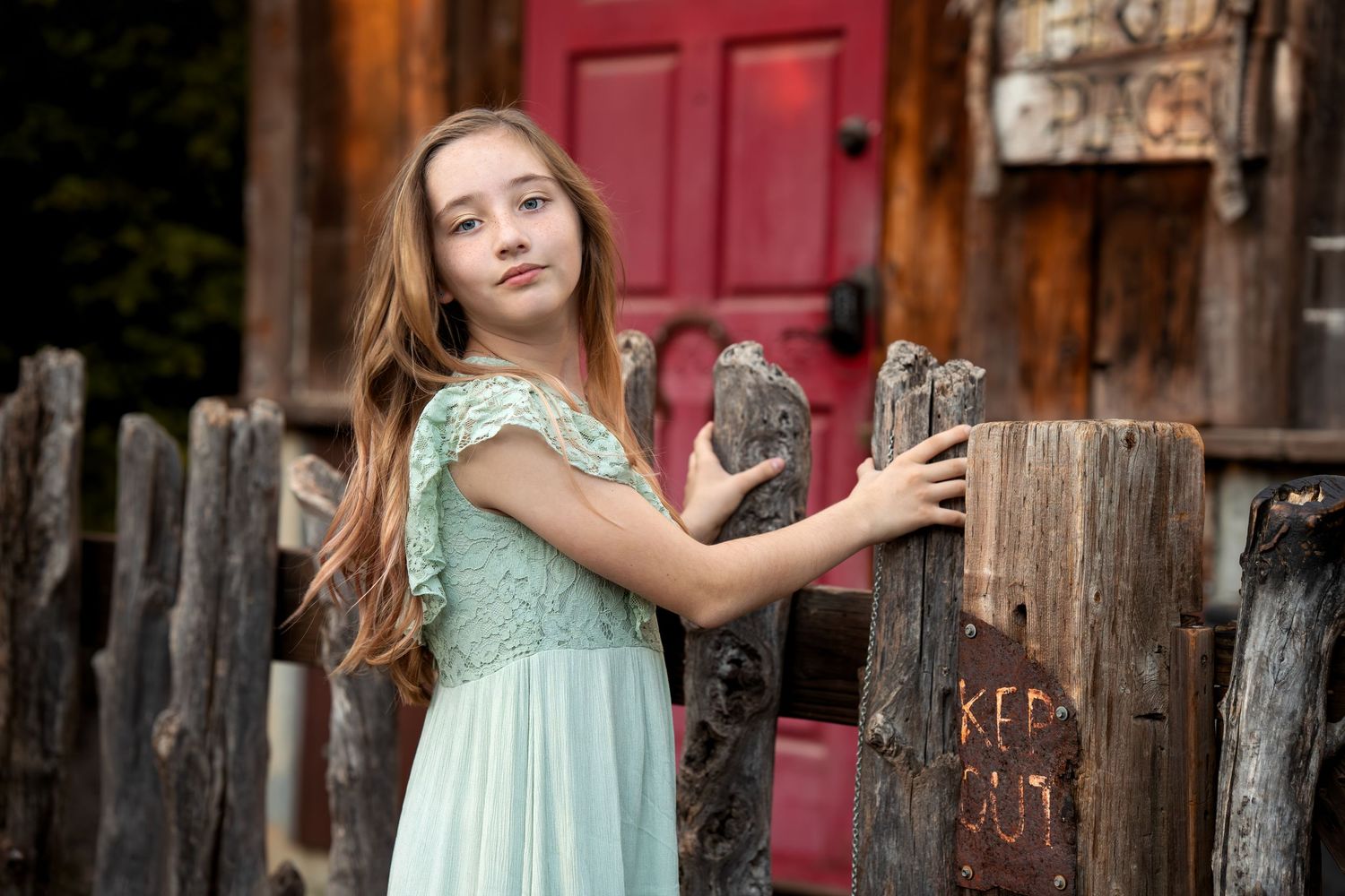 Young girl in a mint green dress poses by a rustic wooden fence with a red door in the background.