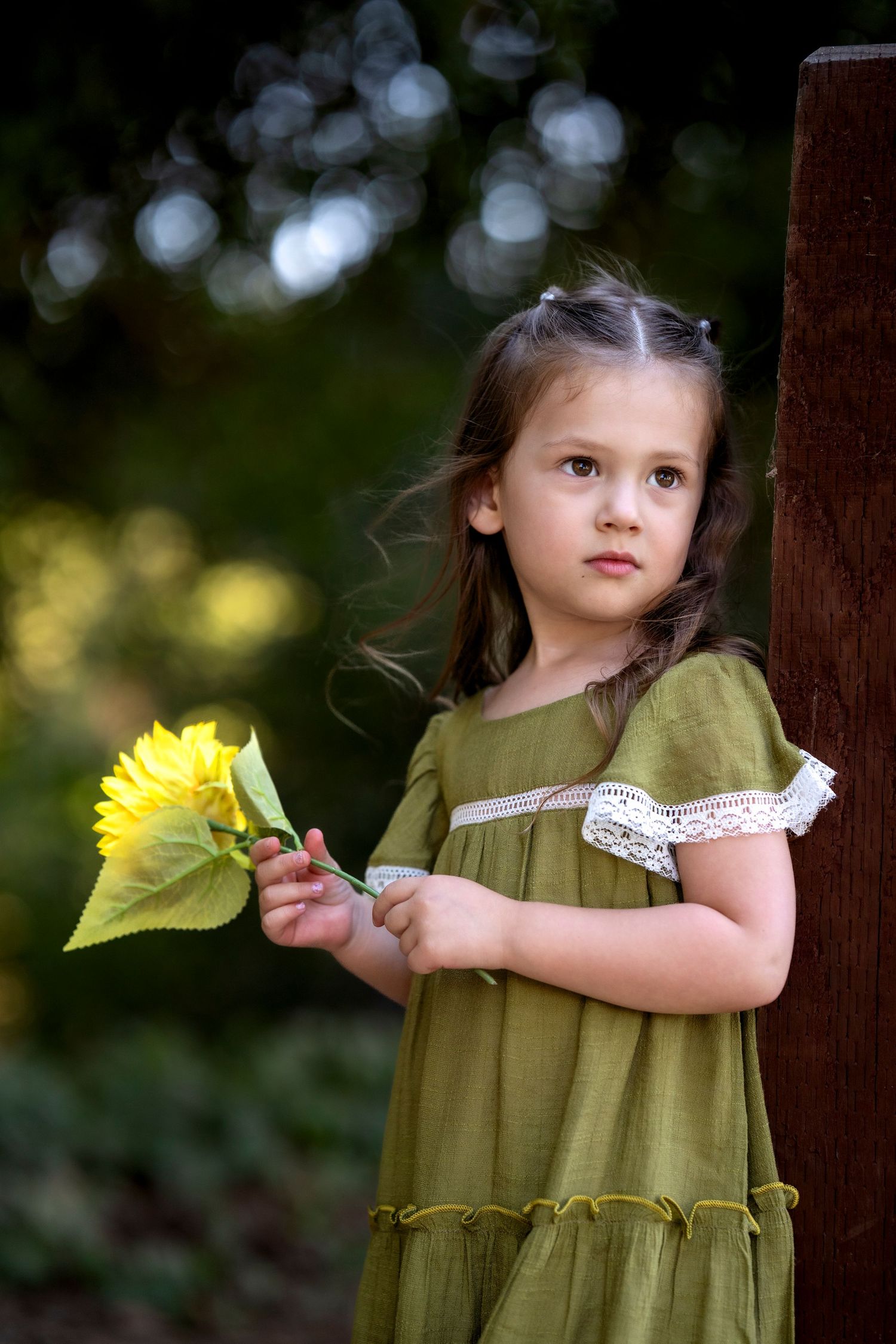 A young child in a green dress holds a yellow daffodil flower against a blurred natural background.