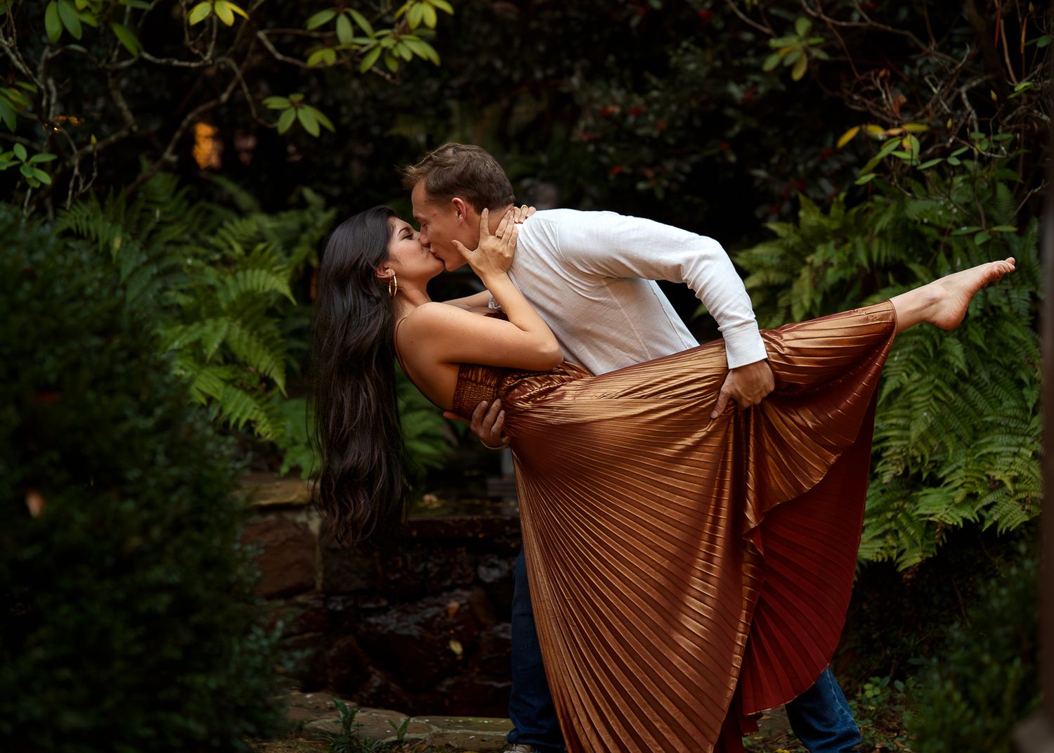 Couple in romantic dance pose outdoors with woman in flowing copper dress against garden backdrop.