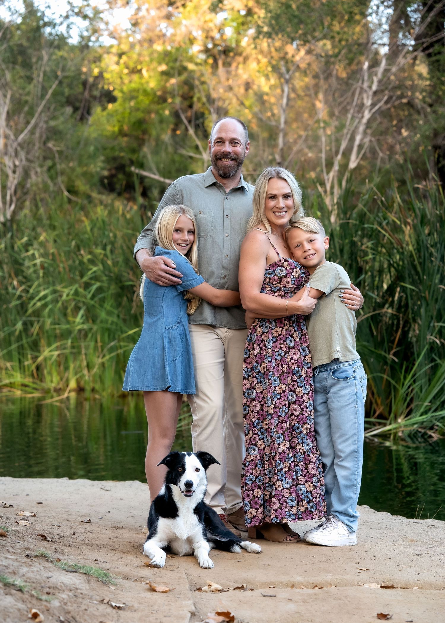 A family of four poses with their black and white border collie dog near a pond surrounded by autumn trees.