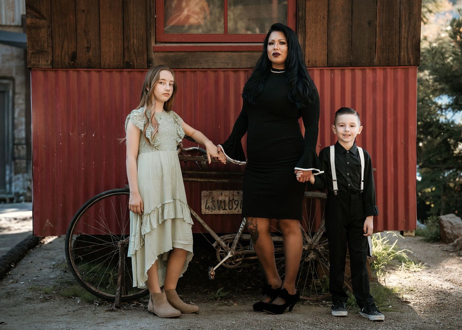 Three people in contrasting black and beige outfits pose together in front of a rustic red wall with an old bicycle.