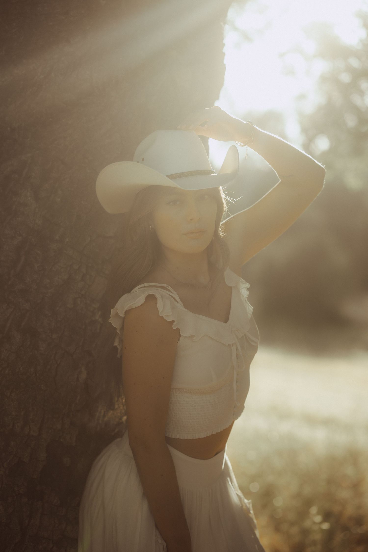 Woman in white cowboy hat and ruffled crop top leans against tree in golden sunlight outdoors.