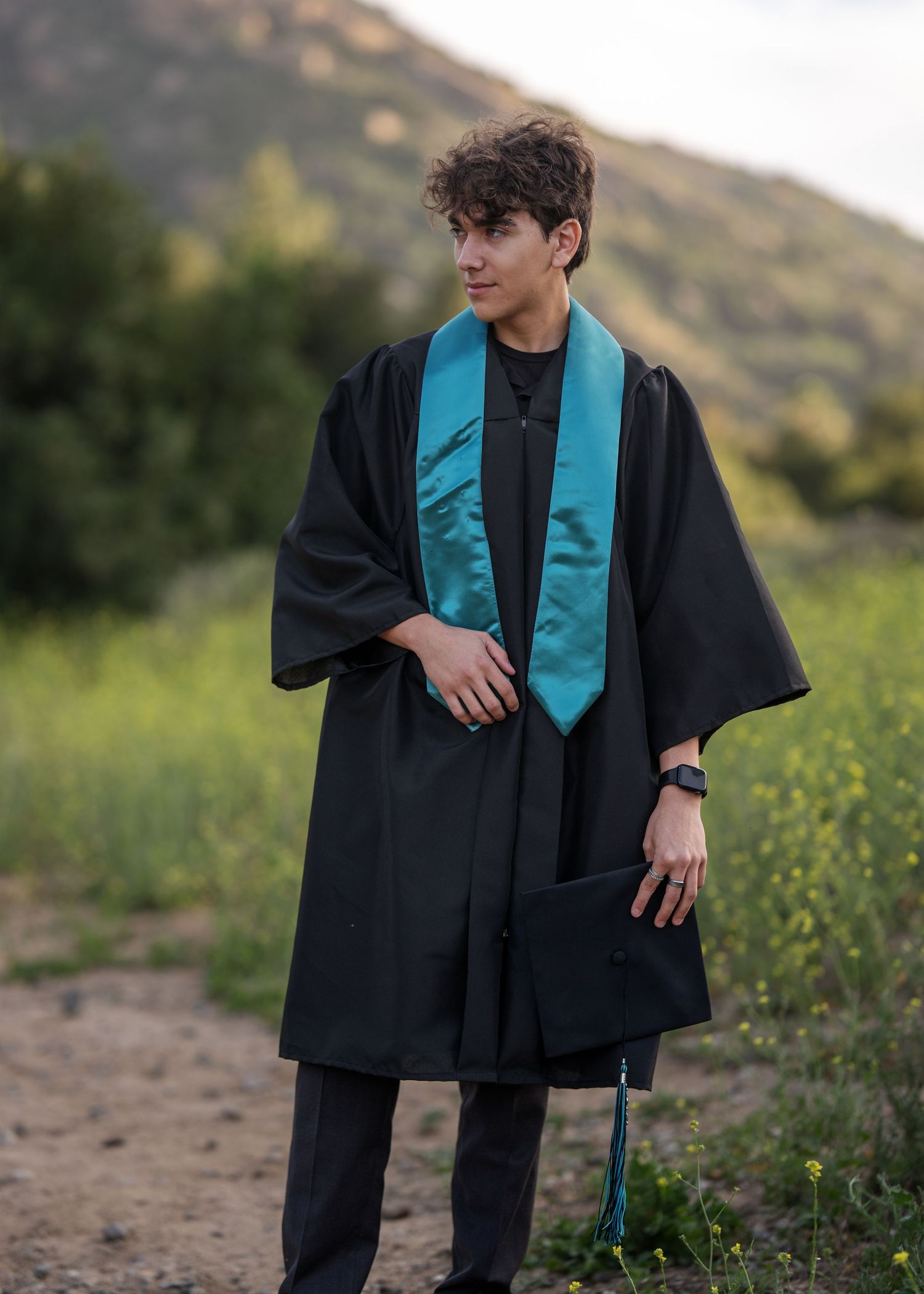 Young man in black graduation gown with teal stole holding cap outdoors on a hillside path.