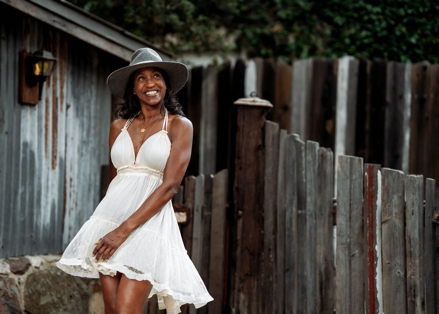 A joyful person in a flowing white dress twirls and dances in front of a rustic wooden fence.