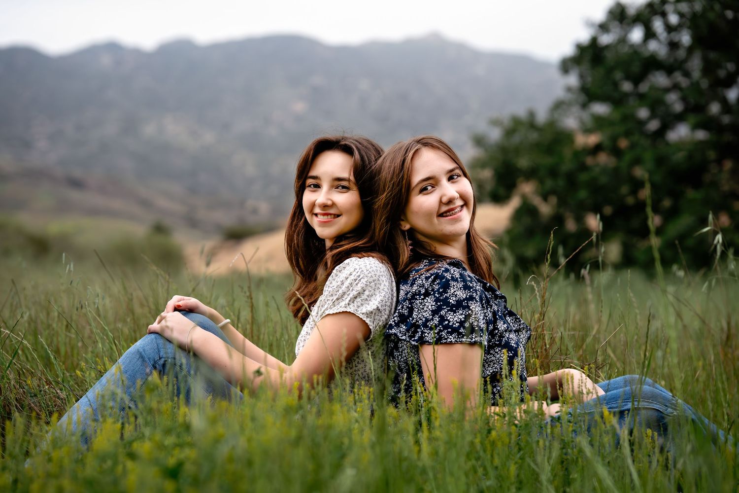 Two friends sit back-to-back laughing in a grassy meadow with mountains in the background.