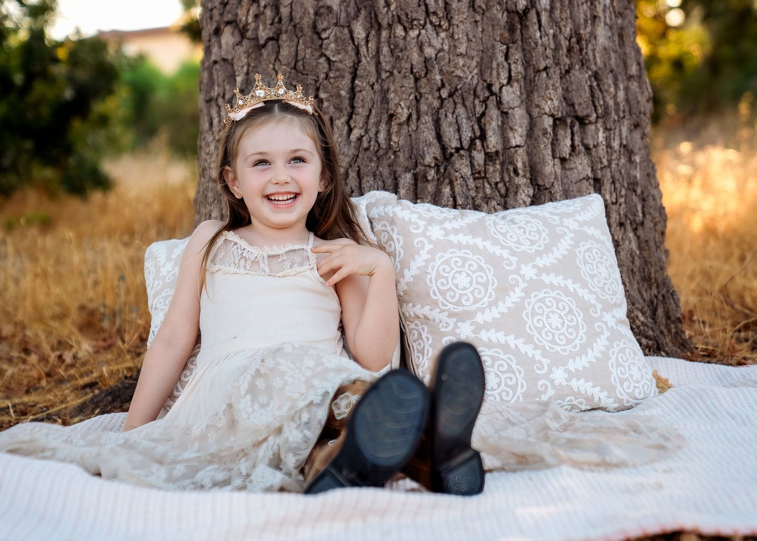 White lace dress and decorative pillows arranged by large tree trunk in outdoor setting.