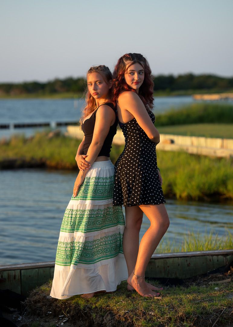 Two young women pose back-to-back near a waterfront, one in a green maxi skirt, the other in a polka dot dress.