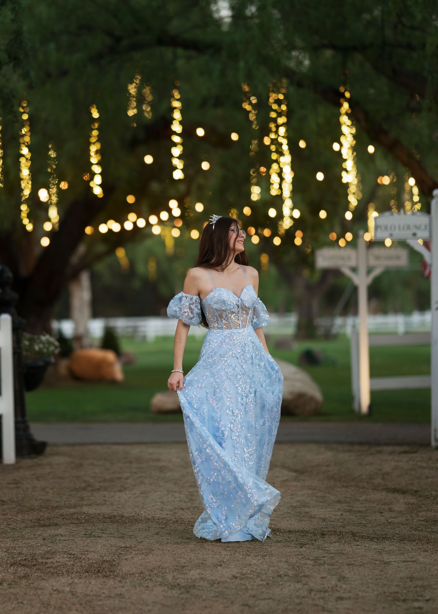 Person in a flowing light blue lace gown walks through an outdoor venue with twinkling string lights in the trees.