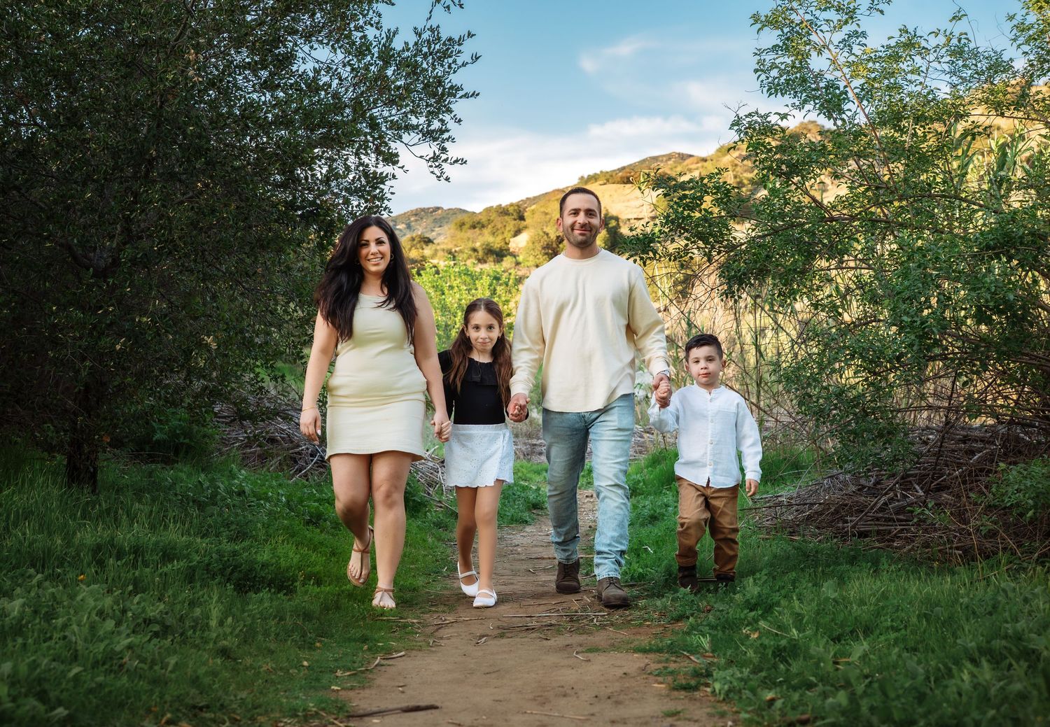 A family walks together on a dirt path through green foliage with golden hills in the background on a sunny day.