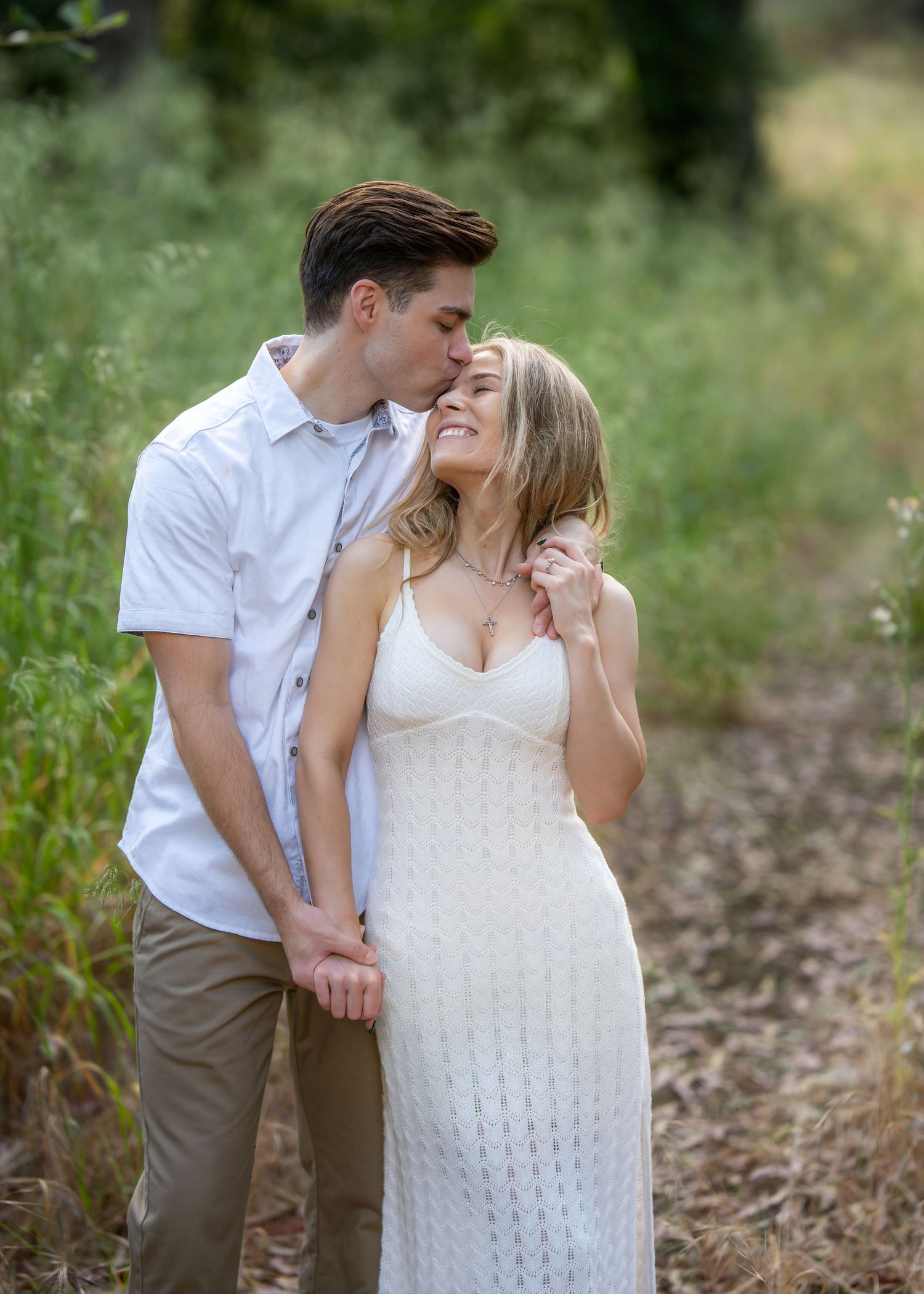 A couple shares a tender moment outdoors, man kissing woman's forehead as she smiles in a white dress.