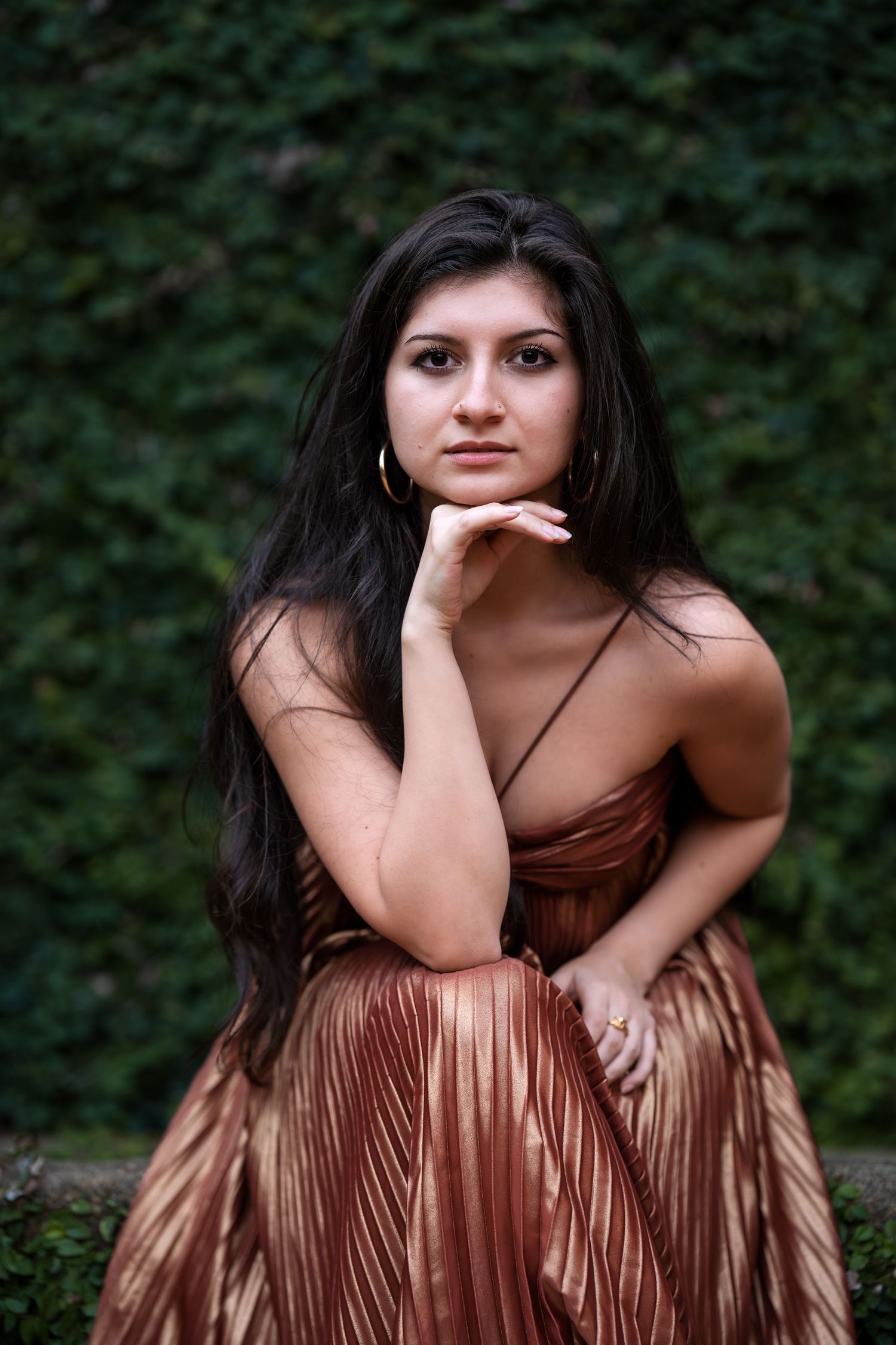 Young woman in bronze pleated dress poses gracefully against lush green ivy wall backdrop.