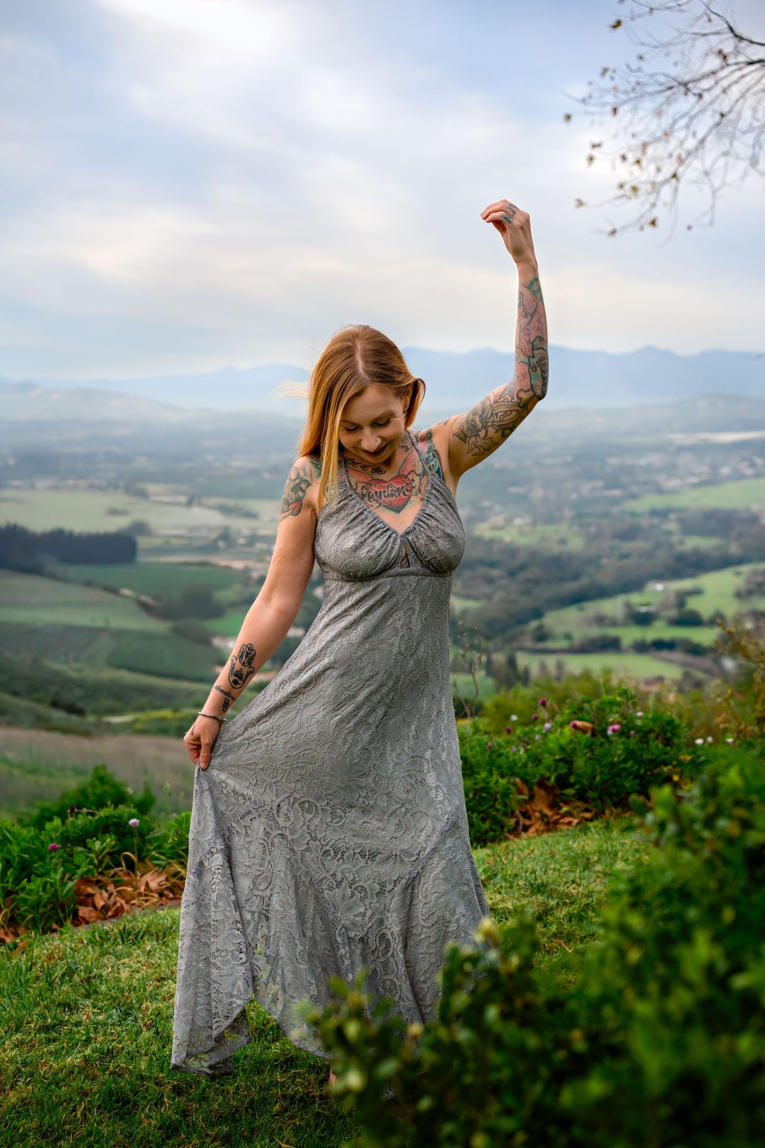 Tattooed woman in gray lace dress dancing outdoors on hilltop with scenic green valley and mountains in background.