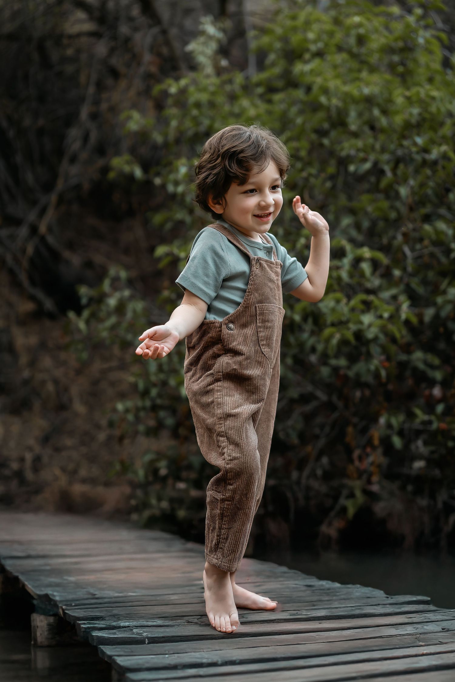 A child in corduroy overalls dances barefoot on a wooden deck.