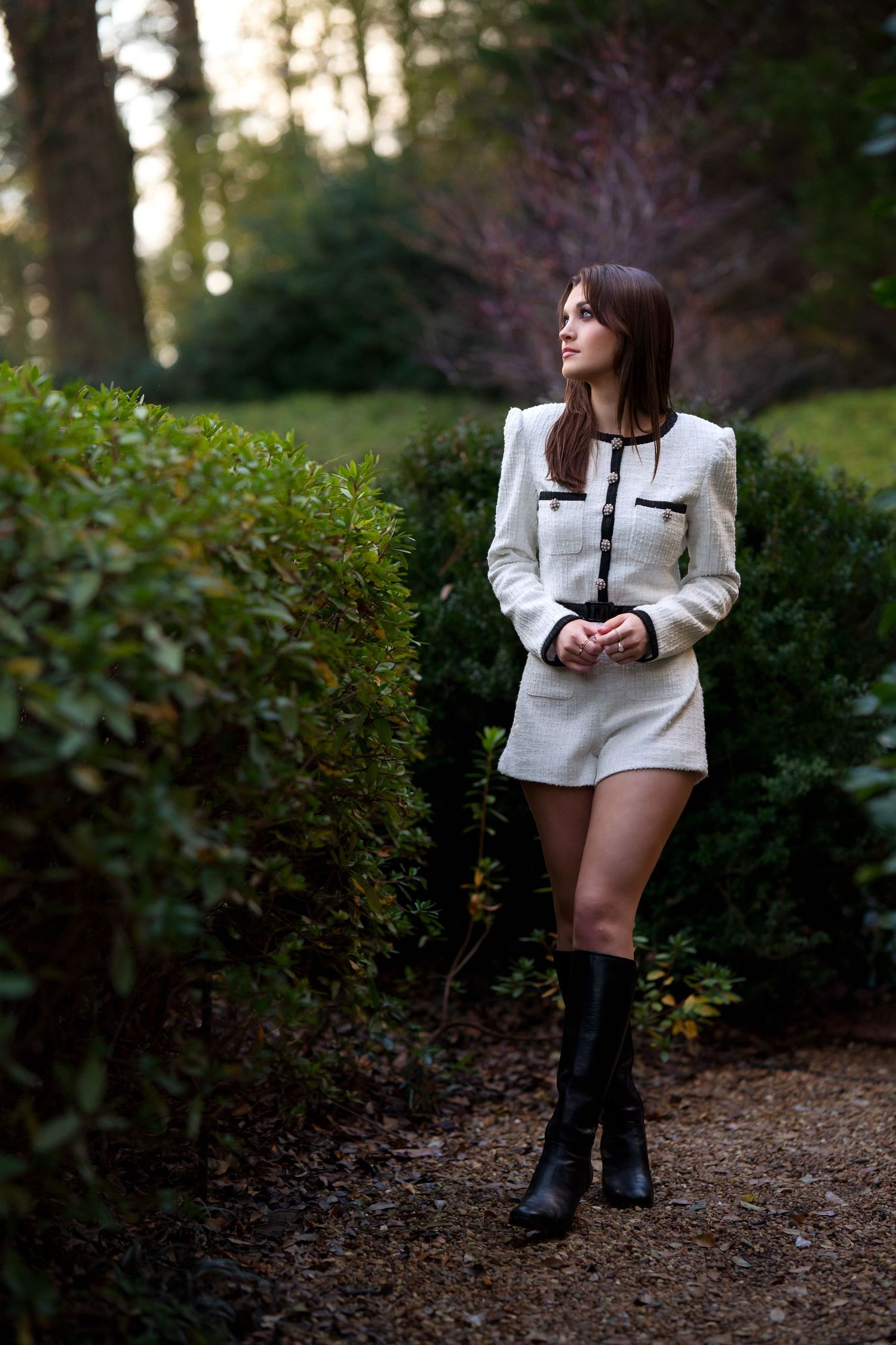 Young woman in white tweed romper and black knee-high boots posing in a lush garden pathway.
