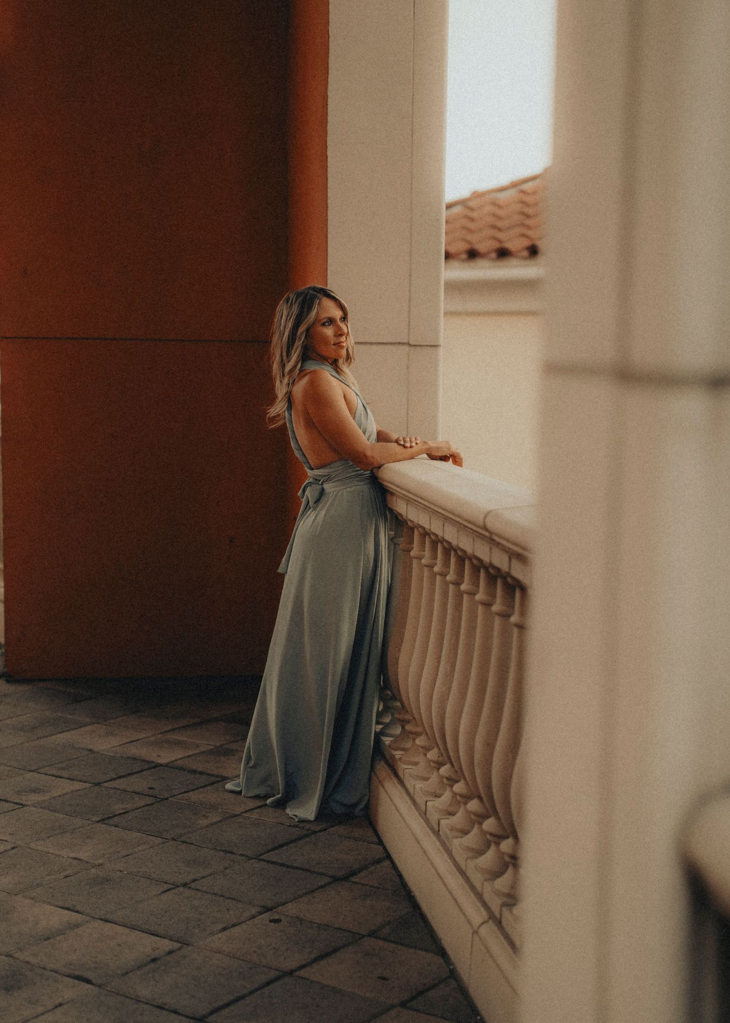 Woman in elegant sage green gown leaning on ornate balustrade of classic Mediterranean-style building.