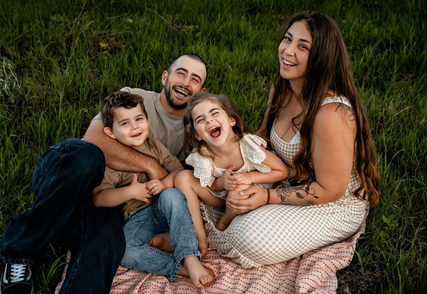 Joyful family portrait with everyone laughing together on picnic blanket in green grass.