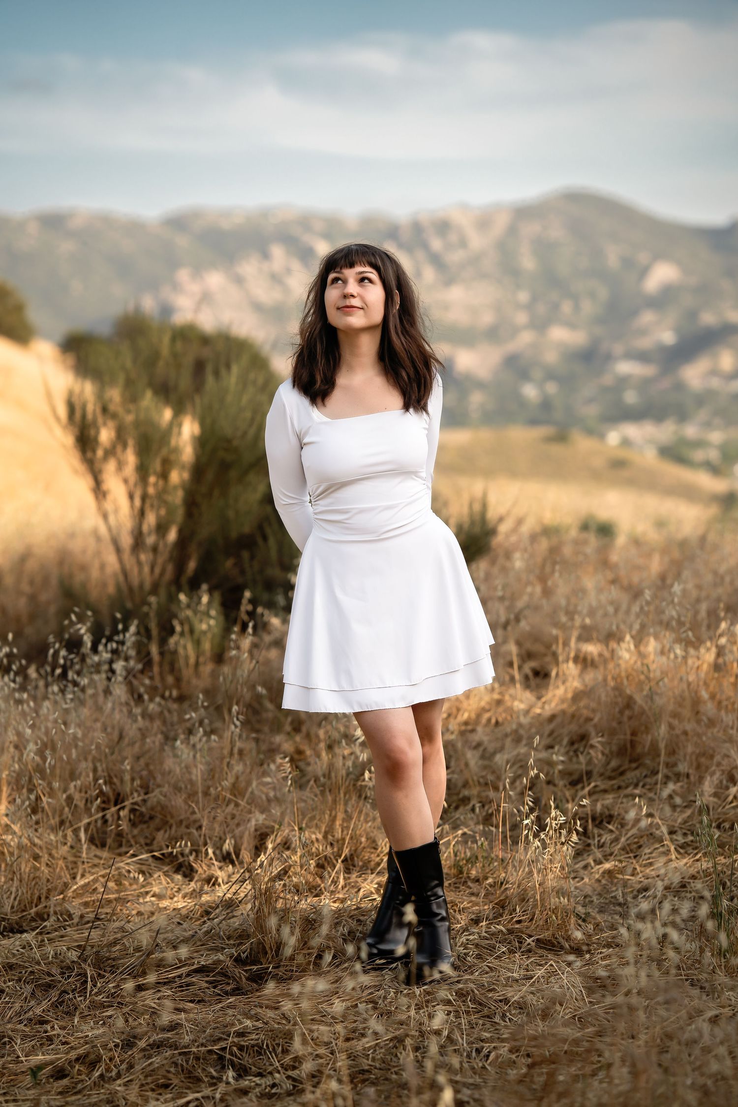 Person in a white dress poses in a field with mountains in the background.