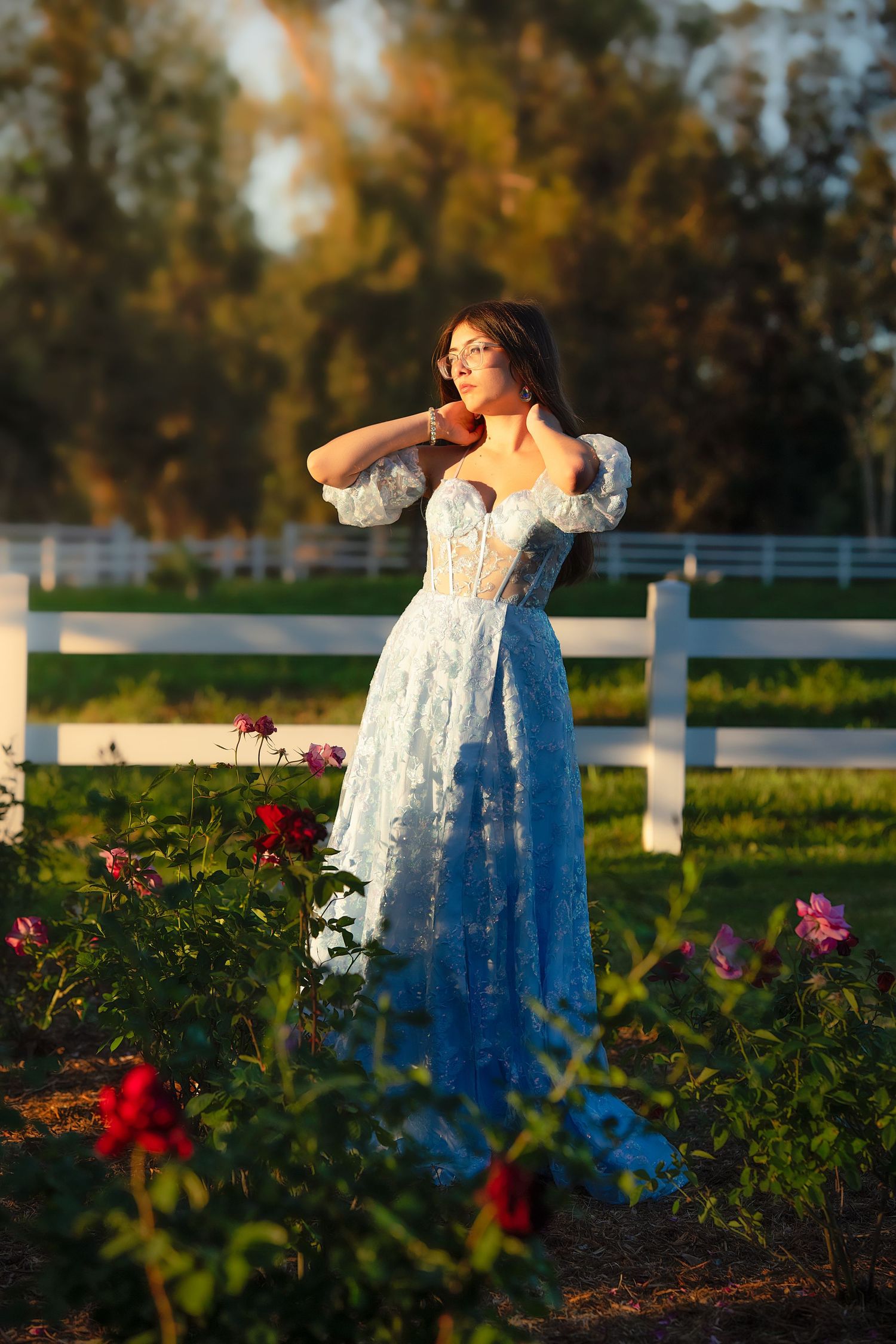 Woman in blue lace gown poses near white fence and roses in golden hour sunlight.