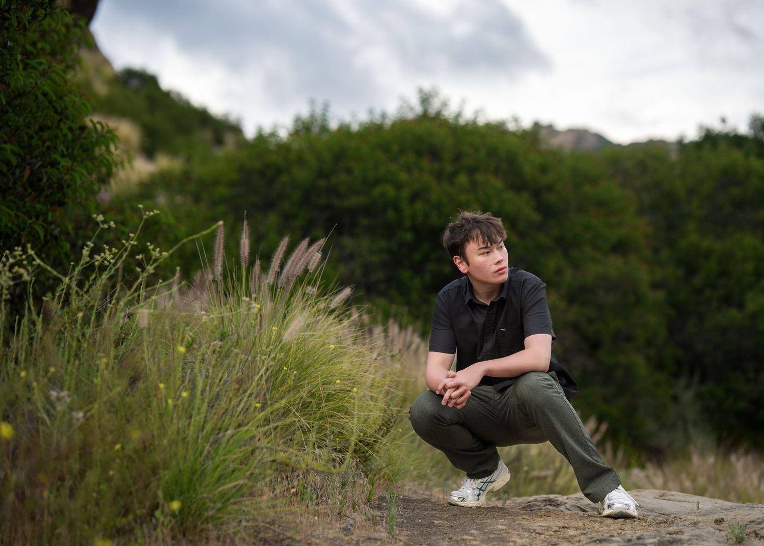 Young Asian man crouching outdoors on a dirt path surrounded by green grass and shrubs under a cloudy sky.