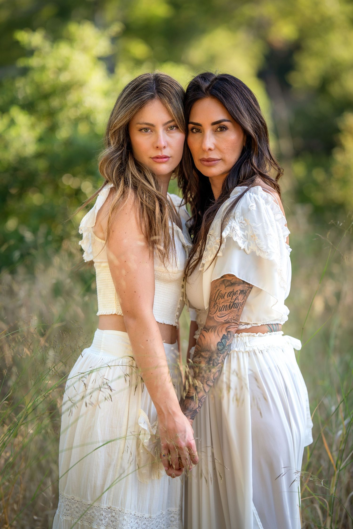 Two women in white boho dresses pose together in a field, one with tattoos, surrounded by lush greenery.