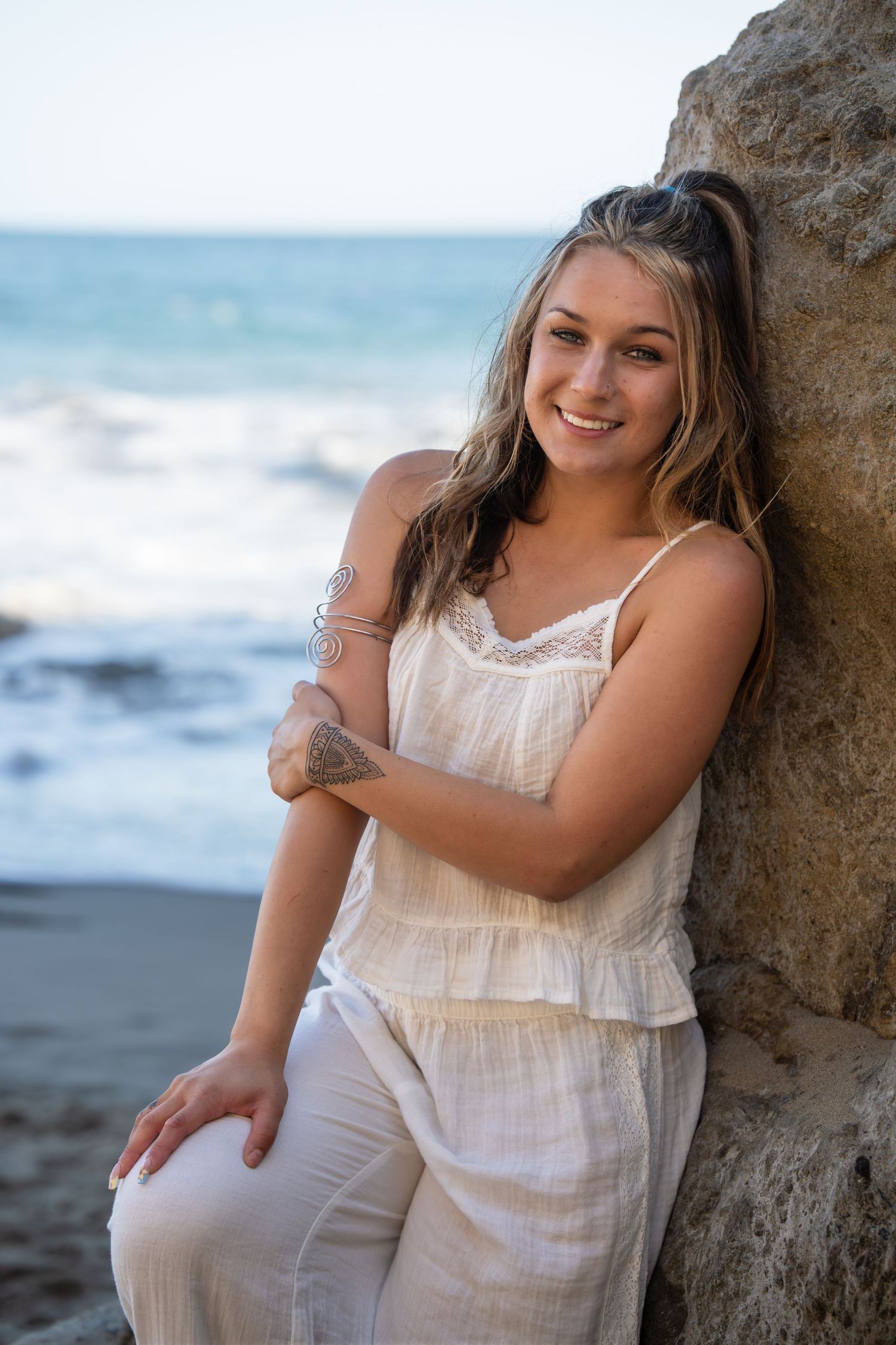 Smiling young woman in white outfit leaning against a rock at the beach with ocean waves in the background.