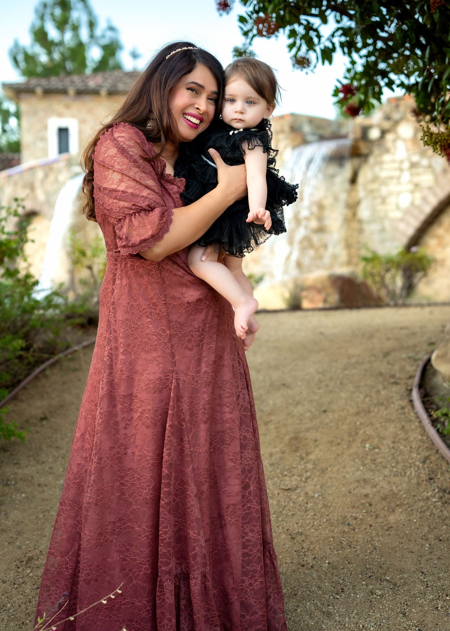 Woman in burgundy dress poses with small child on hillside with stone wall and trees in background.