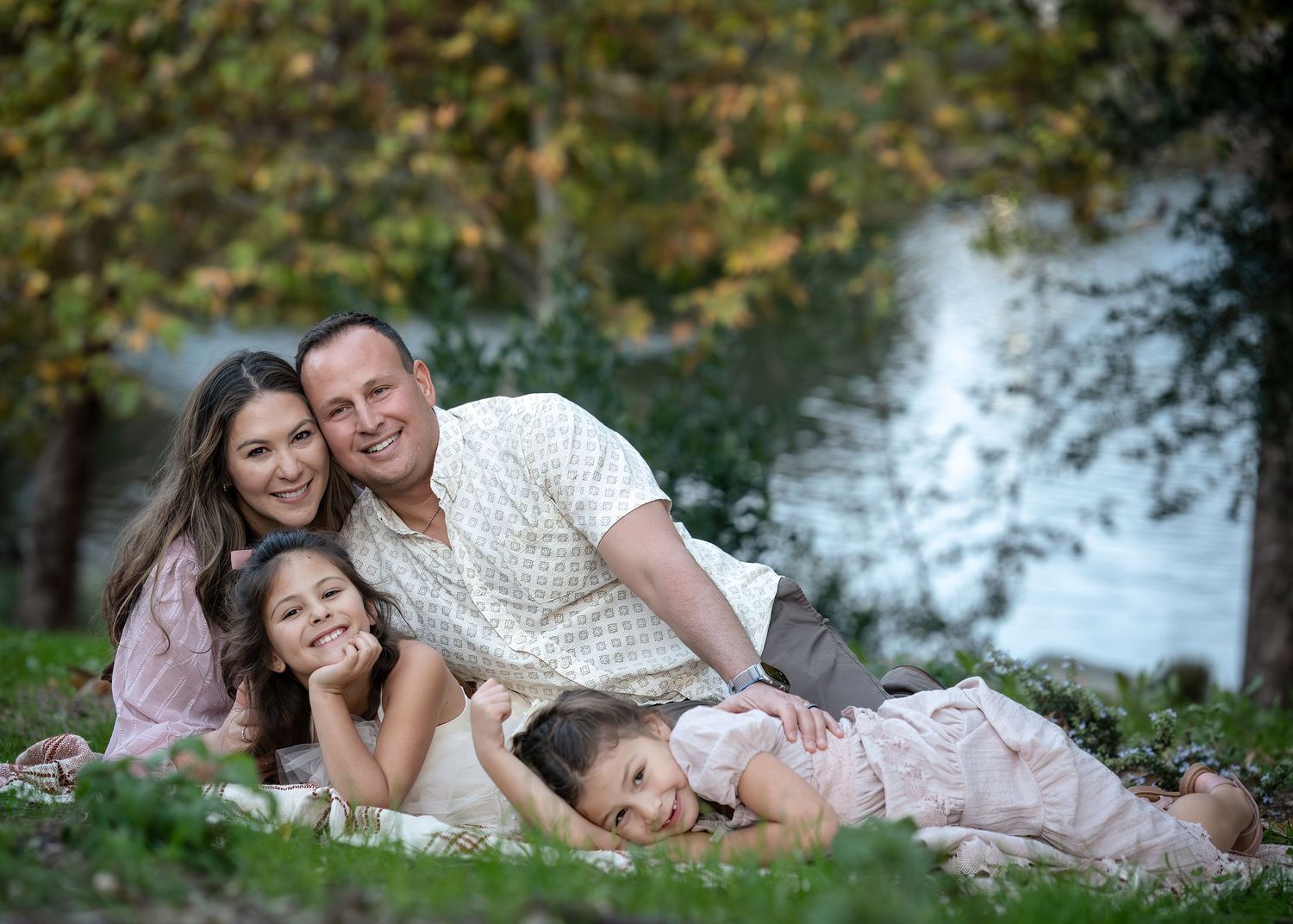 A loving family relaxes together on grass near a peaceful lake during an outdoor photo session.