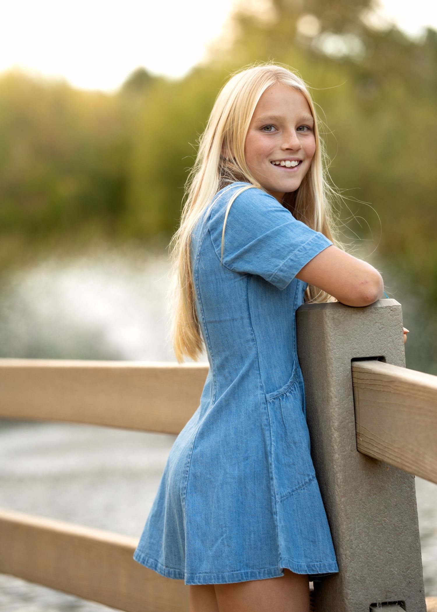 Child in denim dress leaning against wooden railing outdoors during golden hour.