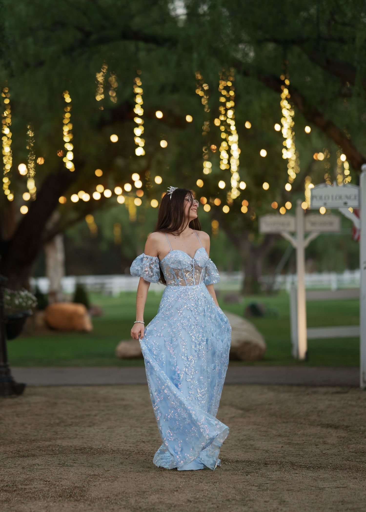 Woman in light blue off-shoulder lace gown standing outdoors under trees with warm string lights at dusk.